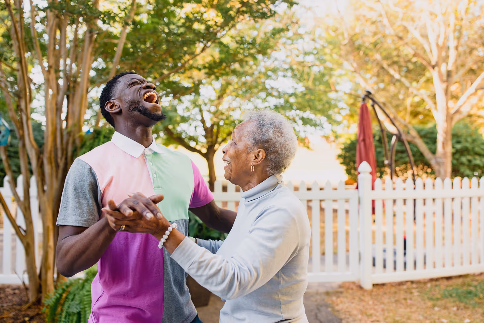 An elderly woman and a younger man are holding hands and laughing together outdoors in a garden area with trees and a white picket fence in the background.