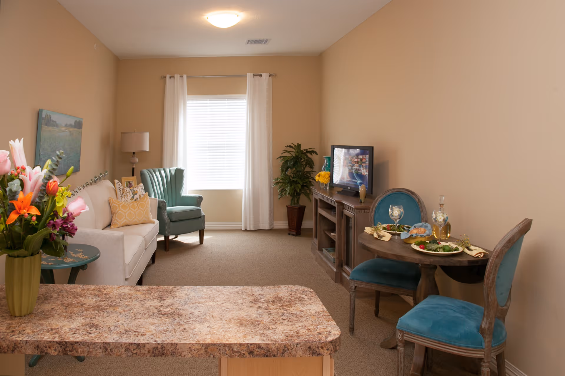 A cozy living room area with beige walls and carpet. The room features a white sofa with decorative pillows, a green armchair, a small round dining table with two blue upholstered chairs, and a TV on a wooden stand. A window with white curtains lets in natural light, and there is a vase of colorful flowers on a countertop in the foreground.