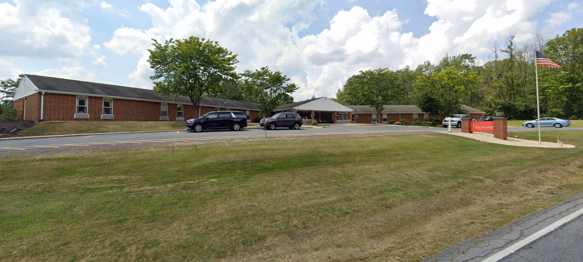 Exterior view of Richland Nursing and Rehabilitation facility showing a single-story brick building with a parking area in front, several cars parked, a flagpole with an American flag, and a sign with the facility's name. The scene includes a grassy lawn and trees in the background under a partly cloudy sky.