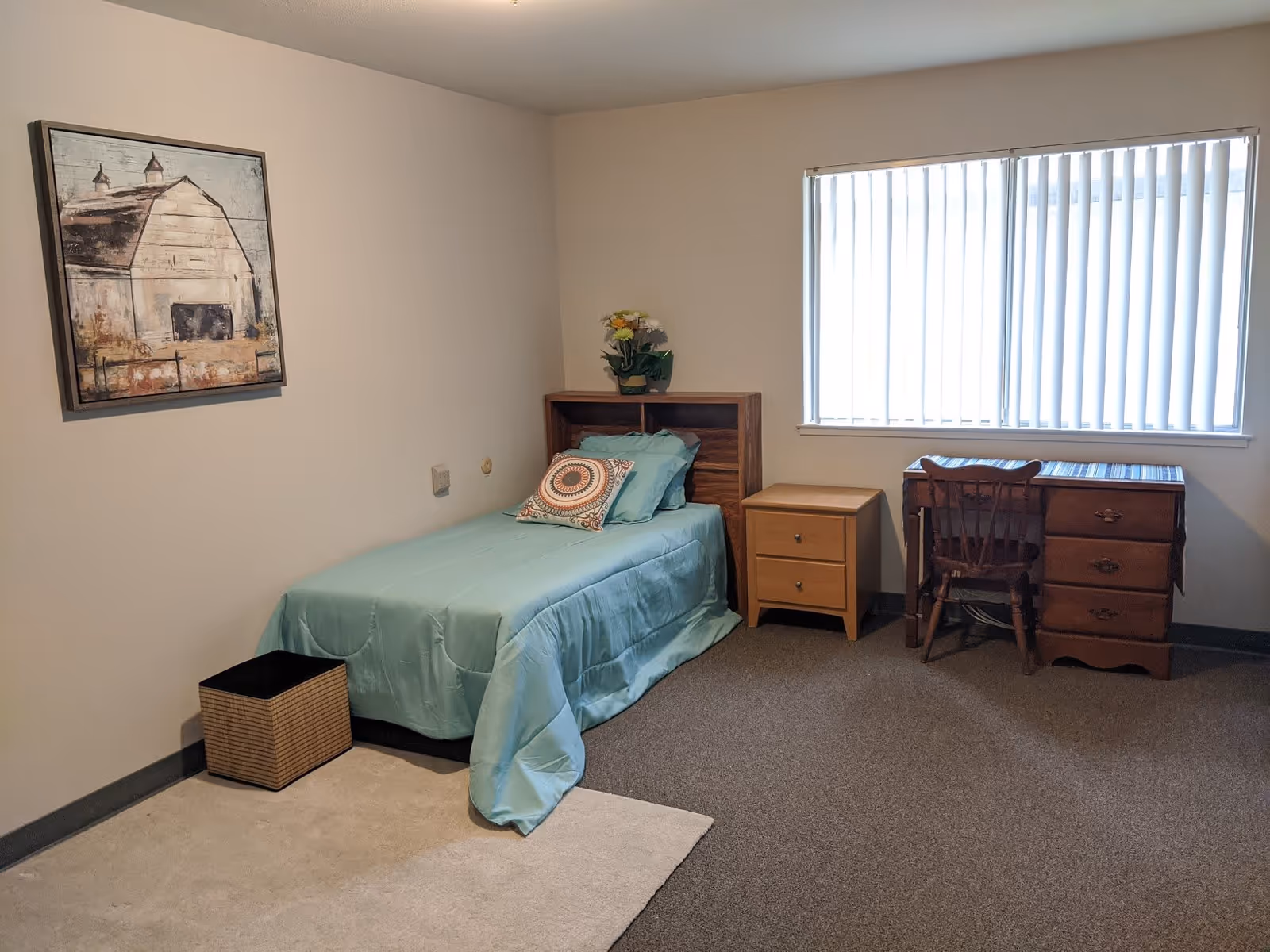 A simple bedroom with a single bed covered in a light blue comforter and a decorative pillow. Next to the bed is a small wooden nightstand with two drawers and a flower pot on top. A wooden desk with a chair is positioned under a window with vertical blinds. A framed painting of a barn hangs on the wall above a small wastebasket on the floor.