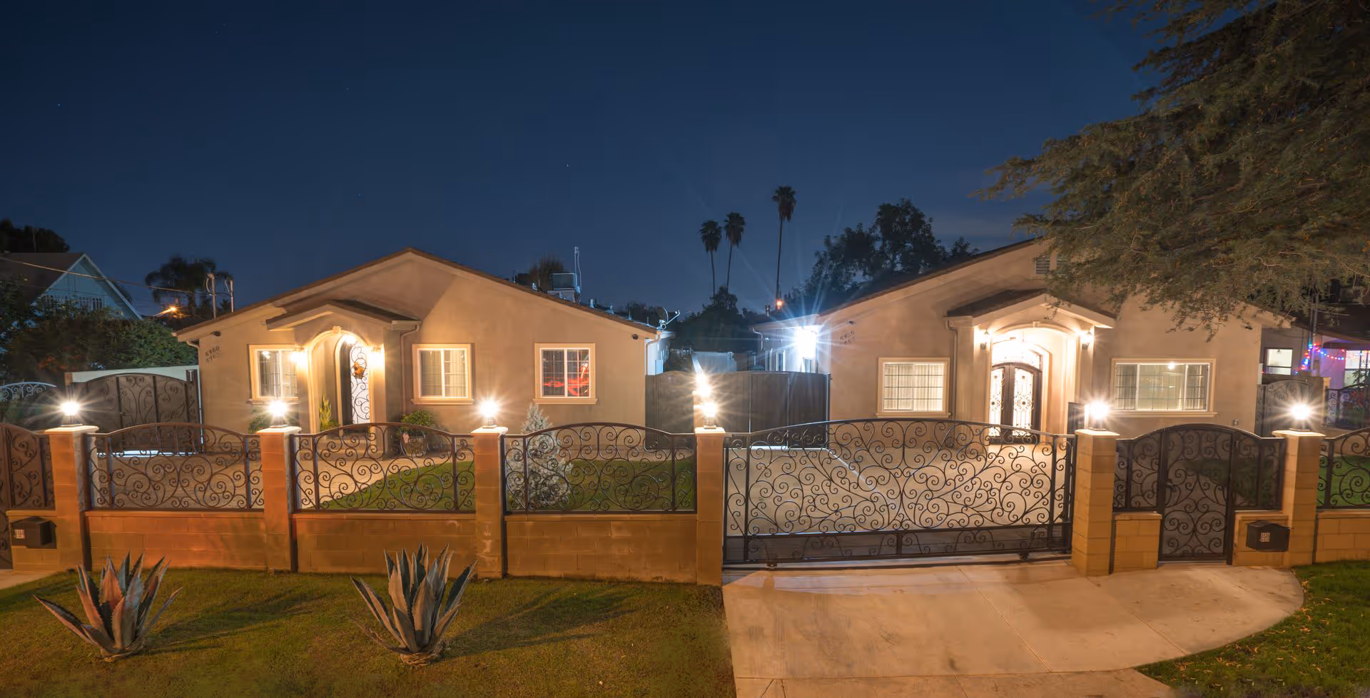 Two single-story houses at night with an ornate wrought-iron front gate and illuminated exterior lights.