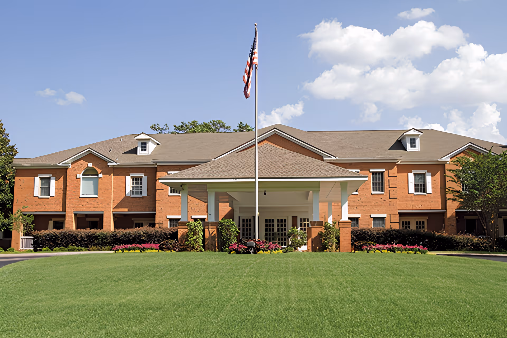 Front exterior view of a large brick building with a covered entrance supported by white columns, an American flag on a flagpole in front, well-maintained green lawn, and flower beds under a partly cloudy blue sky.