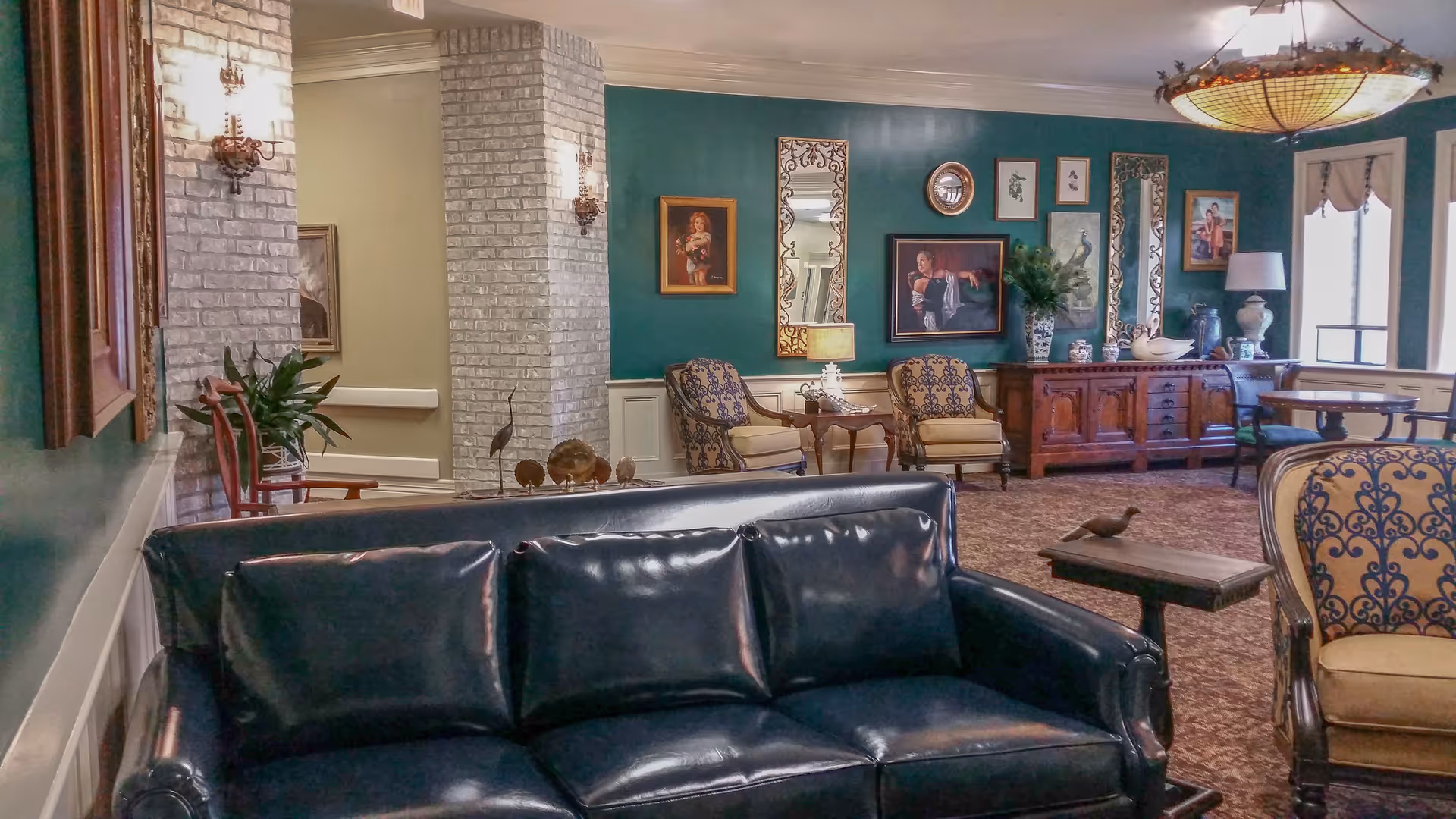 A cozy living room area in a senior living facility with a dark leather sofa in the foreground, patterned armchairs, a wooden side table with a small bird figurine, and a wooden cabinet against a green wall adorned with framed paintings and mirrors. The room has carpeted flooring, wall sconces, and a large window letting in natural light.