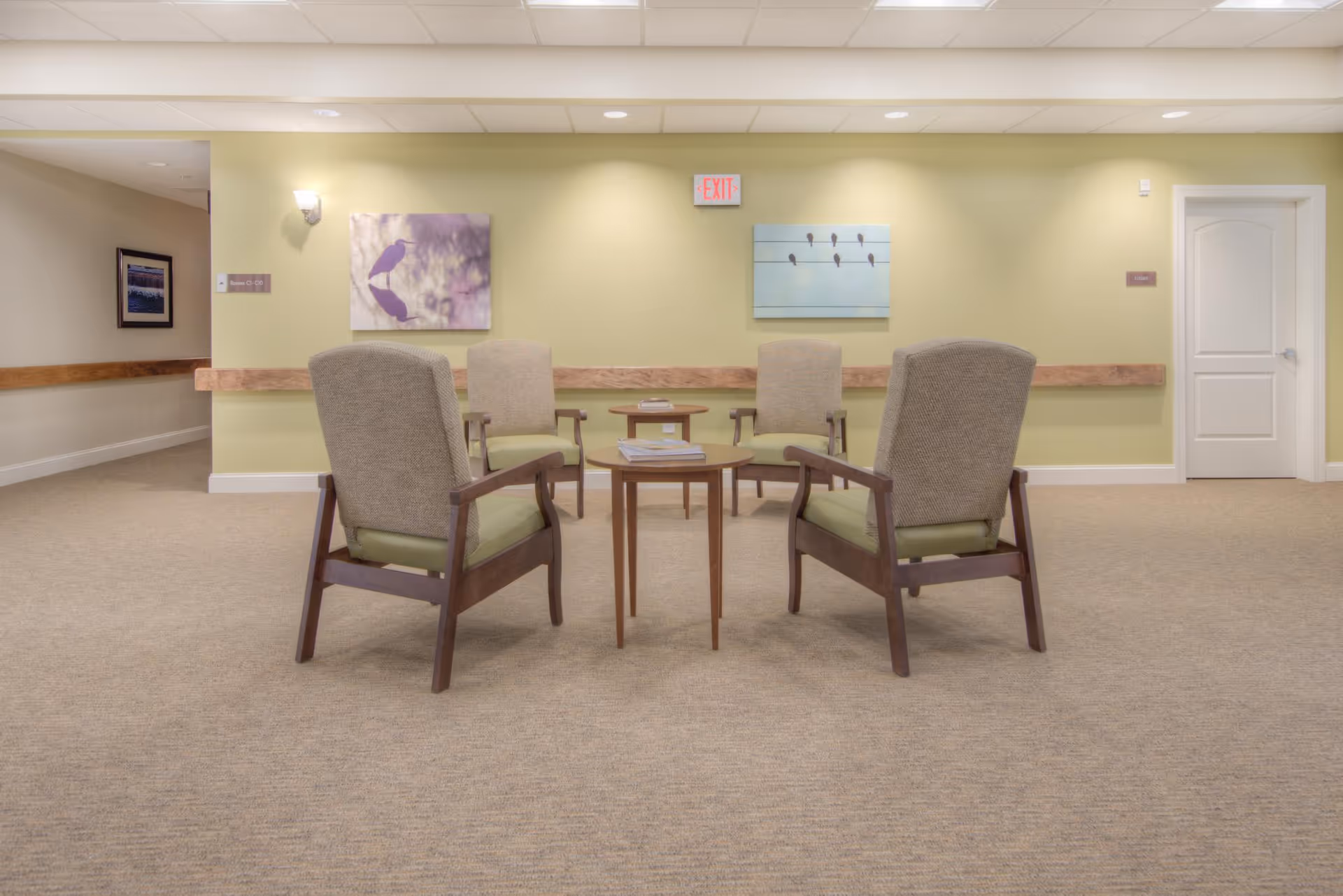 A quiet lounge area with four armchairs arranged around small wooden tables against a pale green wall with artwork and an EXIT sign.