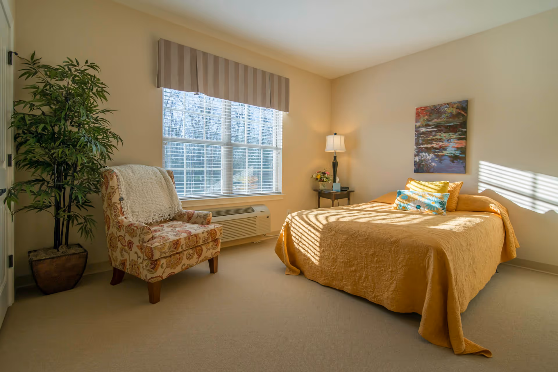 Sunlit furnished bedroom with a bed dressed in a yellow quilt, an armchair, potted plant, bedside table with lamp, and a window with blinds.