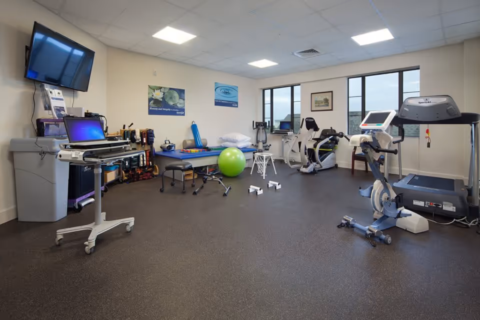 A physical therapy or rehabilitation room with exercise equipment including a treadmill, stationary bike, exercise ball, therapy table, and various small exercise tools. There is a laptop on a mobile stand, a wall-mounted TV, and windows letting in natural light. The walls have motivational posters and a framed picture.