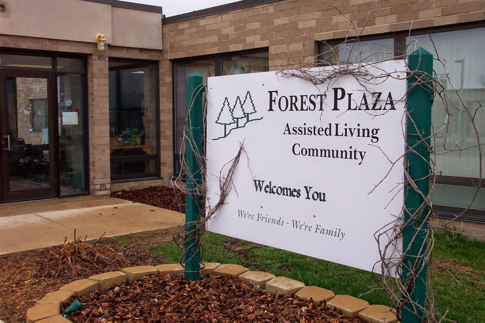 White sign reading "Forest Plaza Assisted Living Community Welcomes You" in front of the brick entrance to an assisted living facility.