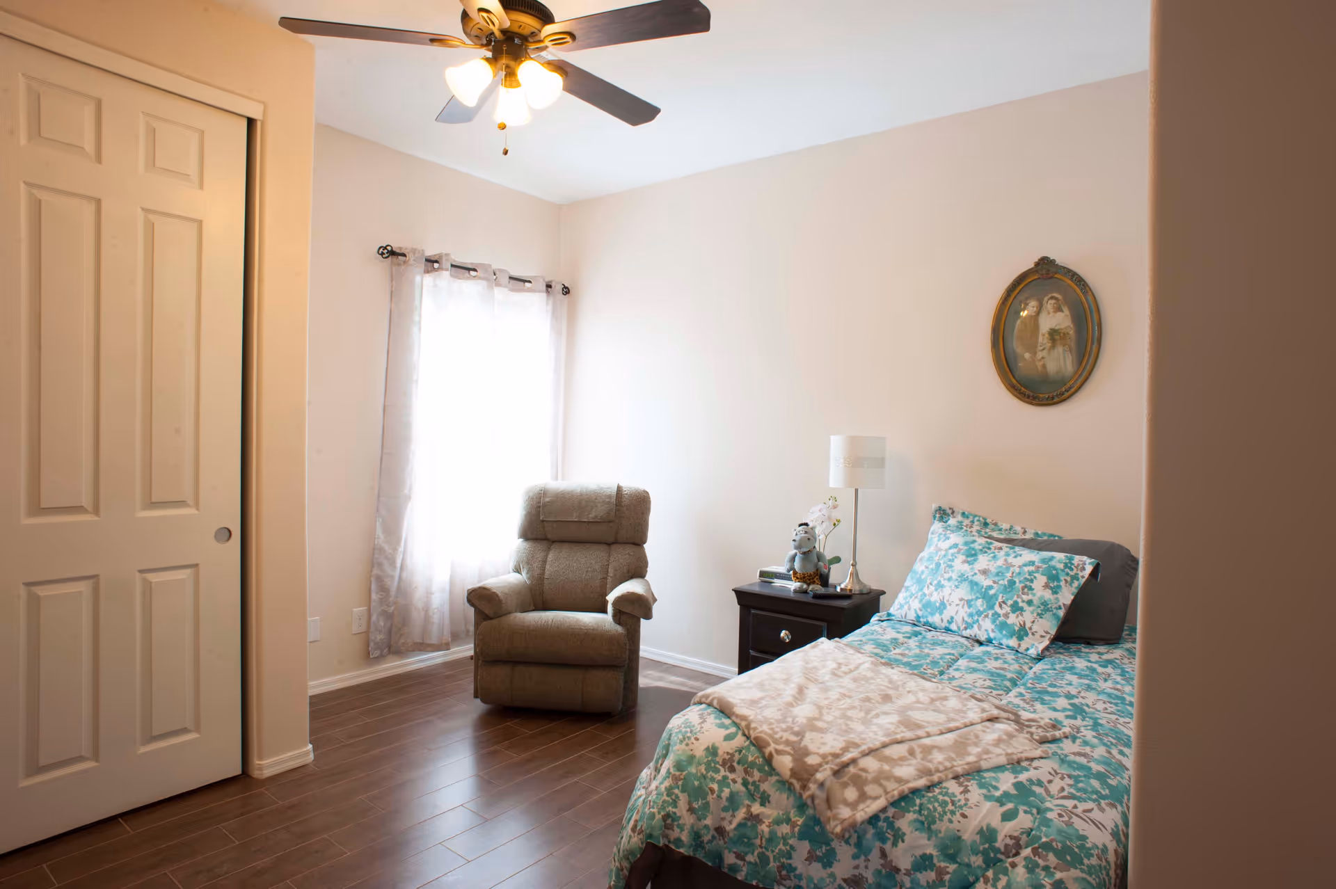 A cozy bedroom with a single bed covered in a floral blue and white comforter and a beige patterned blanket. Next to the bed is a dark wooden nightstand with a lamp, a small plant, and a stuffed animal. A comfortable recliner chair is positioned near a window with sheer curtains. The room has wooden flooring, a ceiling fan with lights, and a framed vintage portrait hanging on the wall.