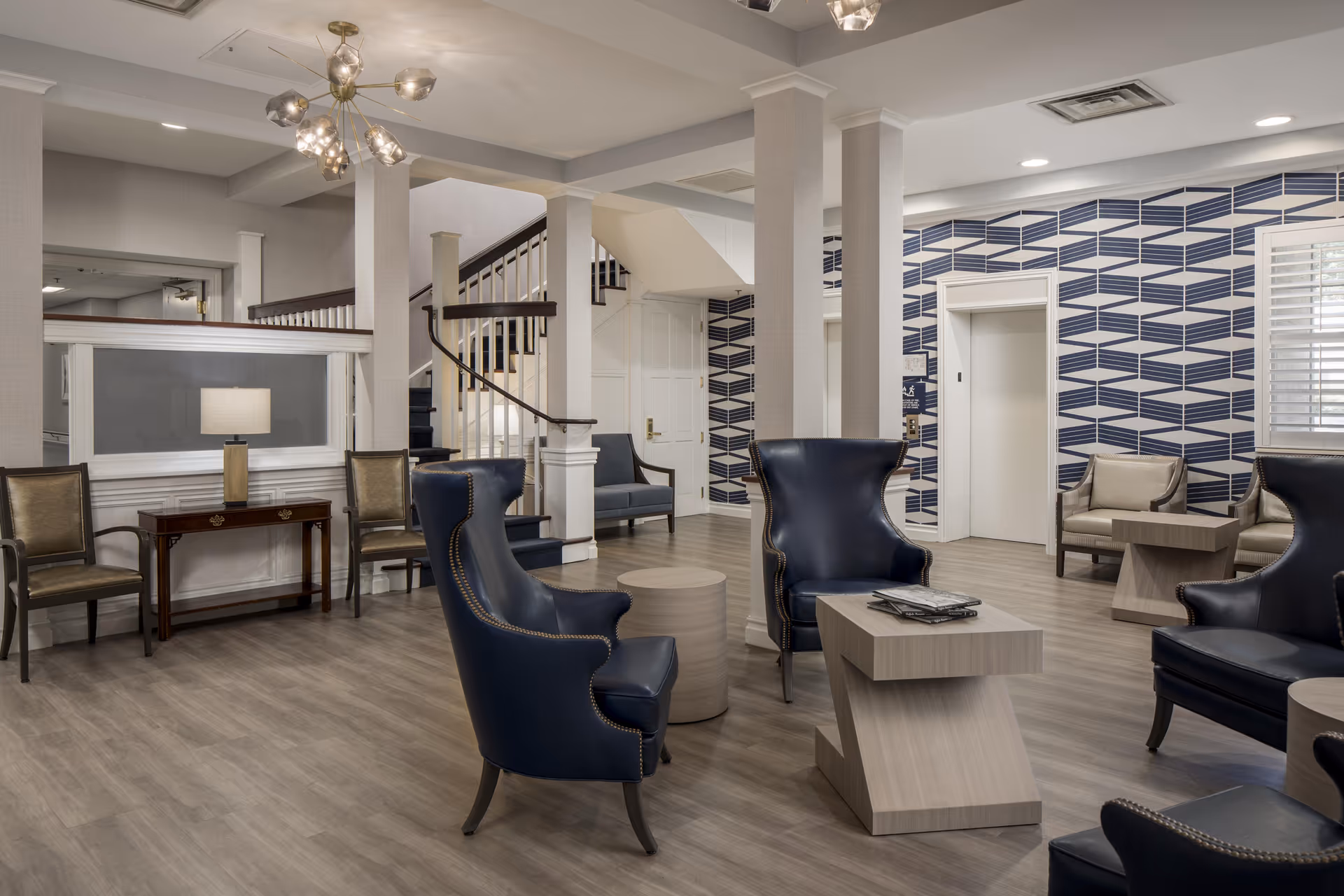 A modern and spacious assisted living facility common area with blue leather armchairs, beige chairs, light wood tables, a staircase with dark wood handrails, and a blue and white geometric patterned accent wall with an elevator door.