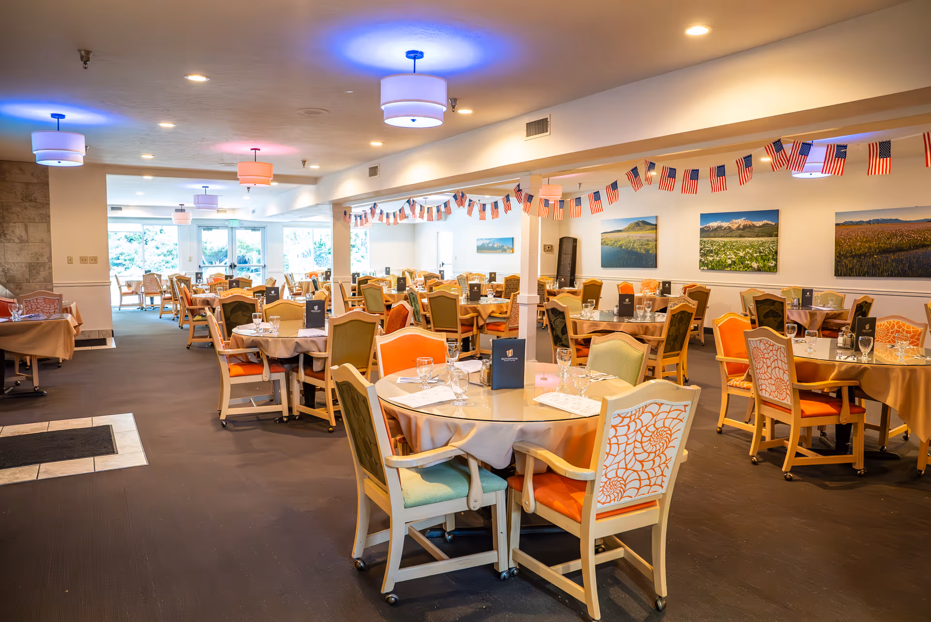 A spacious dining room in a senior living facility with multiple round and rectangular tables covered with beige tablecloths. Each table is set with glasses, napkins, and menus. The chairs are cushioned with various colorful patterns, and American flag garlands hang from the ceiling. The room is well-lit with ceiling lights and has large windows allowing natural light to enter. Landscape paintings decorate the walls.