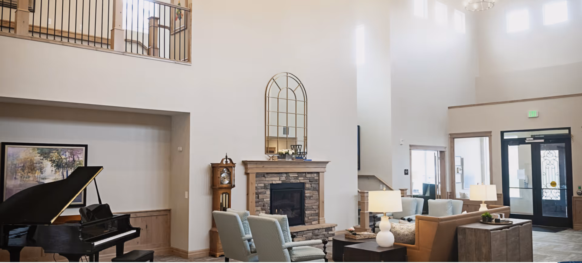 A spacious and well-lit assisted living facility common area with high ceilings, featuring a grand piano, a stone fireplace with a decorative mirror above it, a grandfather clock, comfortable armchairs, sofas, and table lamps. Large windows and glass doors allow natural light to fill the room.