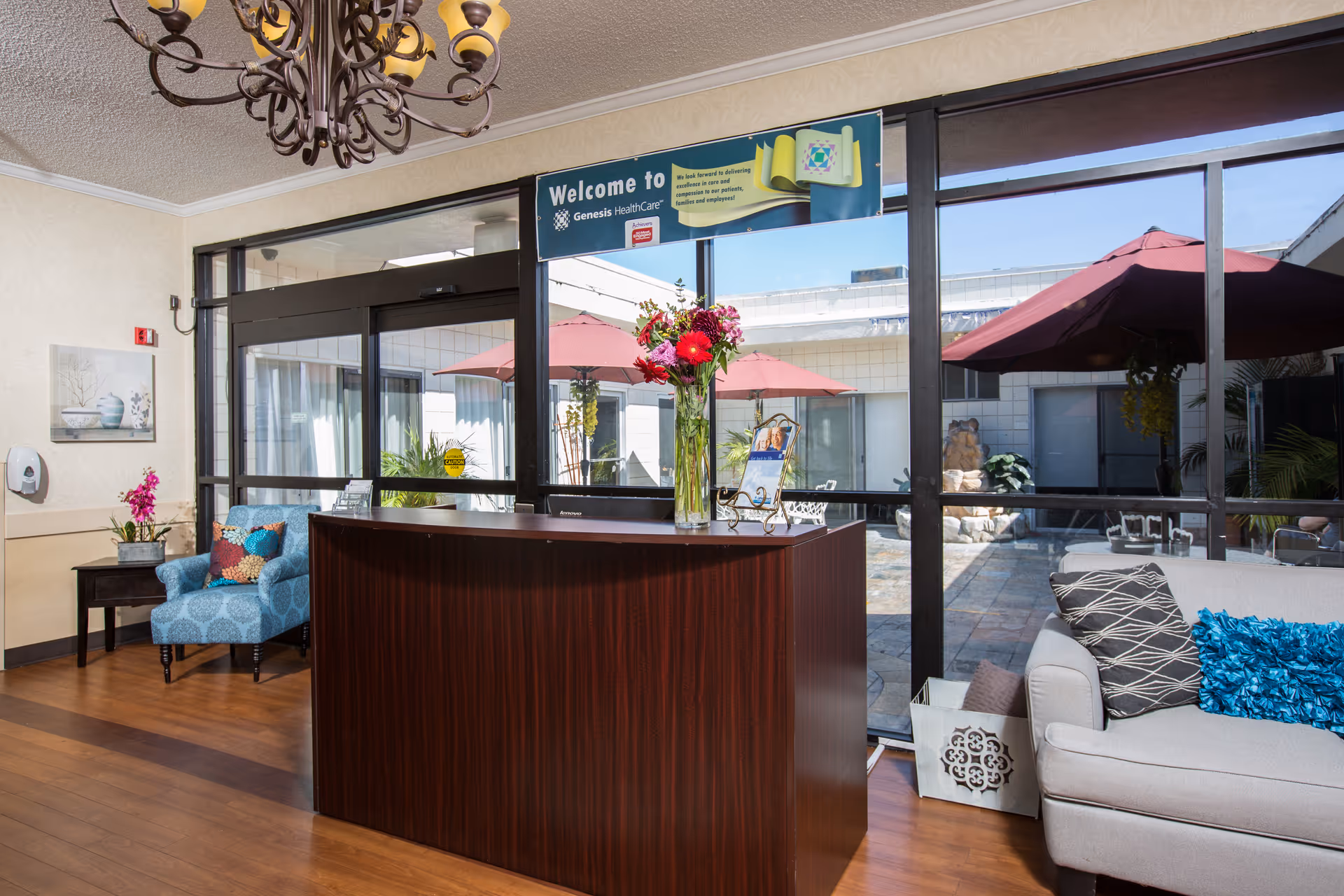 Reception area with a dark wood front desk adorned with a vase of flowers and a small framed sign. Behind the desk is a large window showing an outdoor patio with red umbrellas and seating. To the left, there is a blue armchair with colorful pillows and a small side table with a plant. To the right, a light gray couch with decorative pillows is visible. A chandelier hangs from the ceiling.