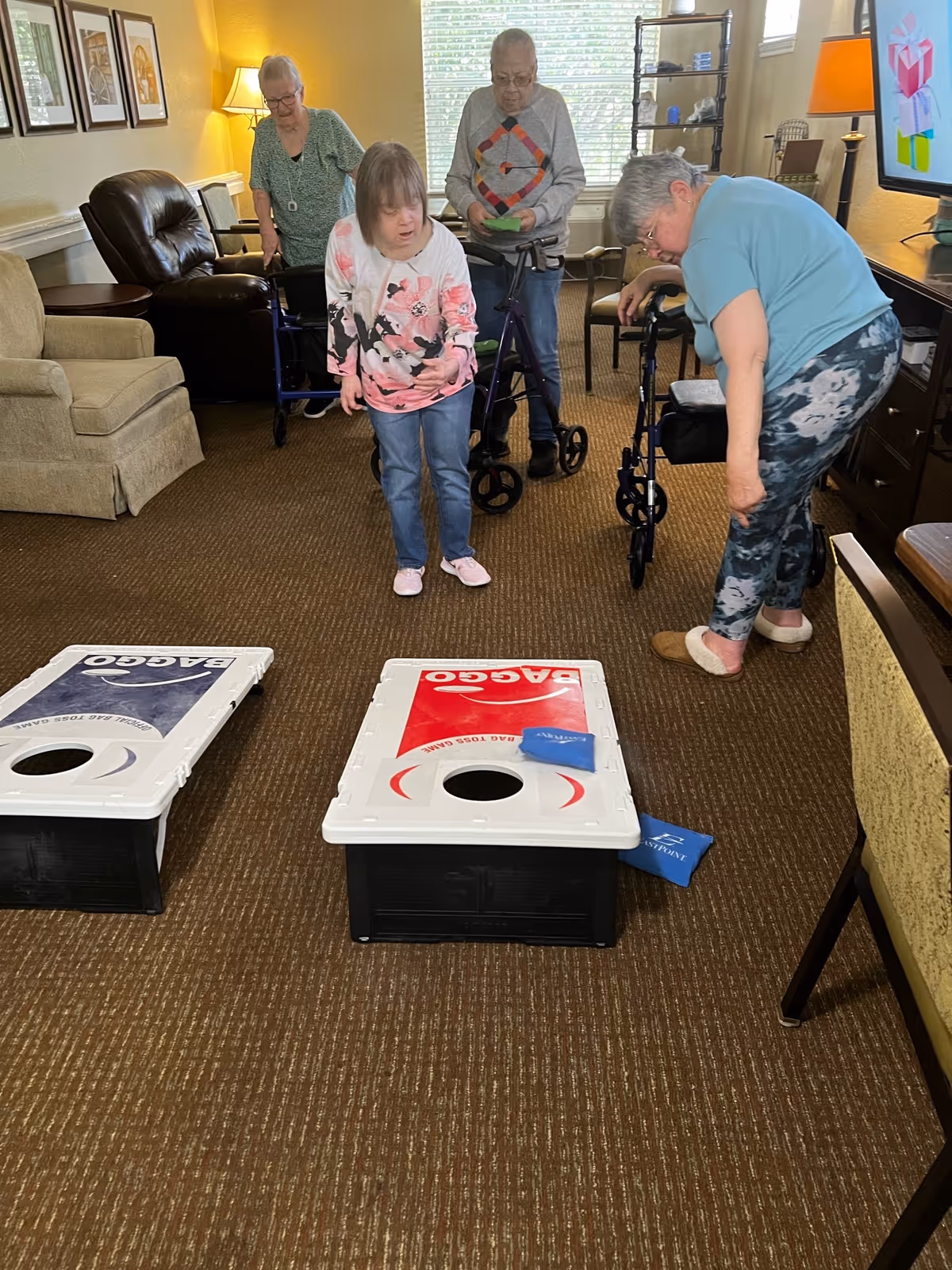Four elderly individuals in a living room area playing a game of cornhole with two cornhole boards placed on the carpeted floor. Two of the individuals are using walkers, and one person is bending down to pick up a bean bag. The room is furnished with armchairs, a lamp, framed pictures on the wall, and a window with blinds in the background.
