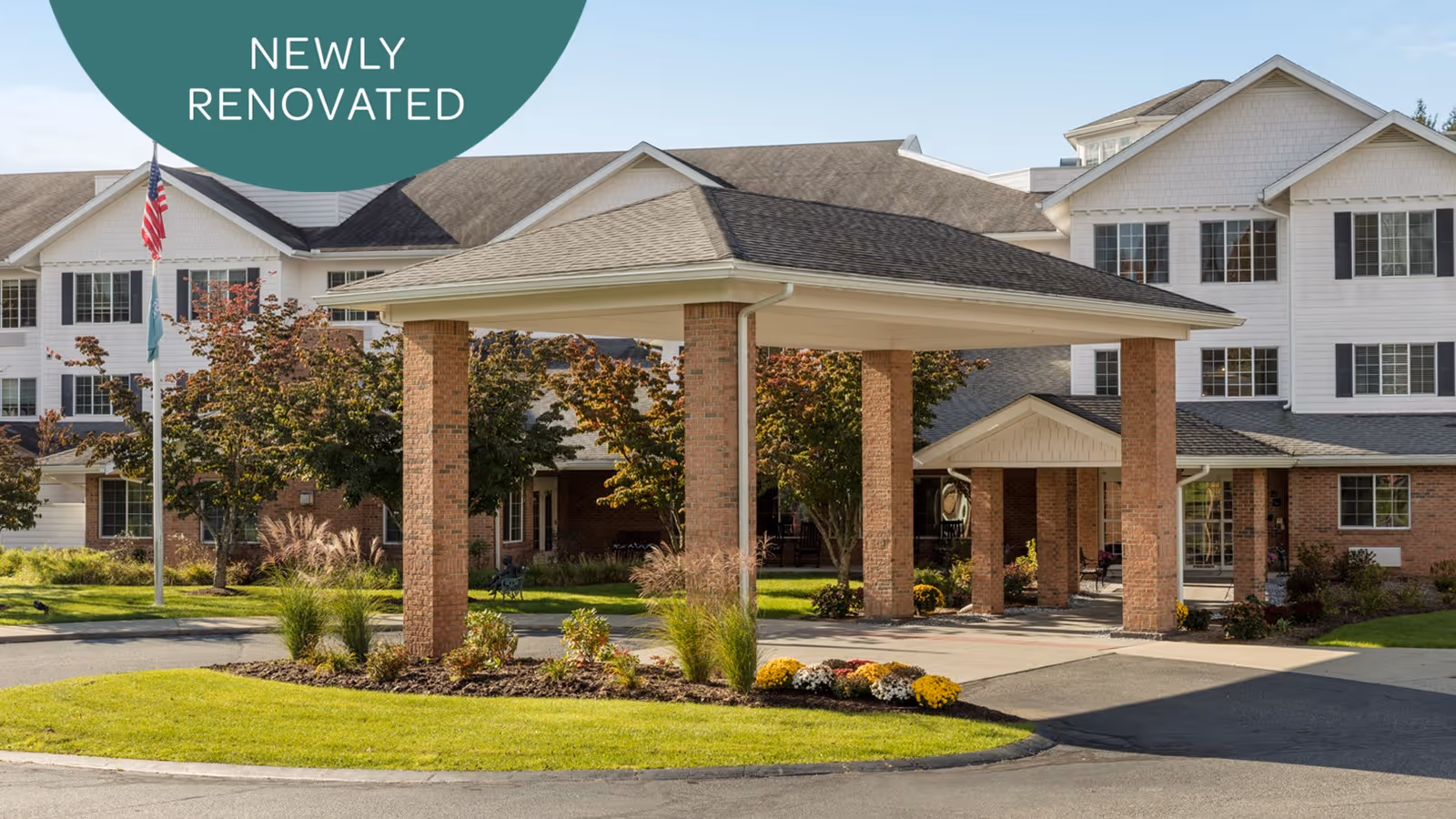 Exterior view of a newly renovated senior living facility with a covered entrance supported by brick pillars, surrounded by landscaped greenery and flowers, with an American flag on a flagpole to the left.
