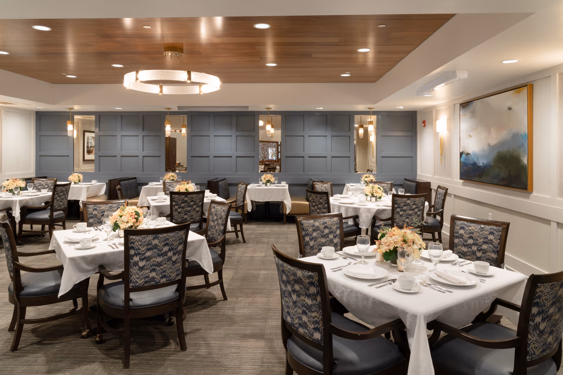 A well-lit dining room with multiple tables covered in white tablecloths, each set with plates, cups, glasses, and silverware. Floral centerpieces adorn each table. The room features patterned chairs, a wooden ceiling section with recessed lighting, and a large abstract painting on the wall.