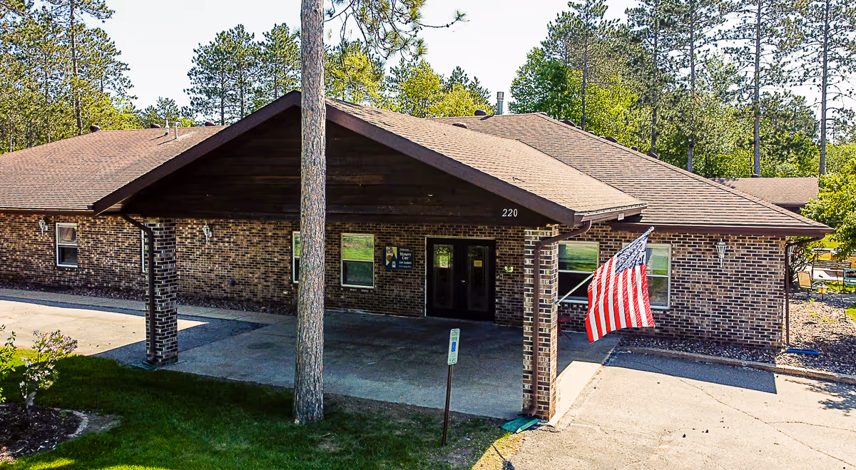 Exterior view of a single-story brick building with a covered entrance supported by brick columns. An American flag is mounted near the entrance, and there are trees and greenery surrounding the building. The building number 220 is visible above the entrance.