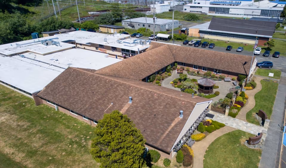 Aerial view of a senior living facility with a large brown roof forming a U-shape around a landscaped courtyard featuring a gazebo, benches, and walking paths. Surrounding the building are green lawns, shrubs, and a parking area with several cars. Additional buildings and train tracks are visible in the background.