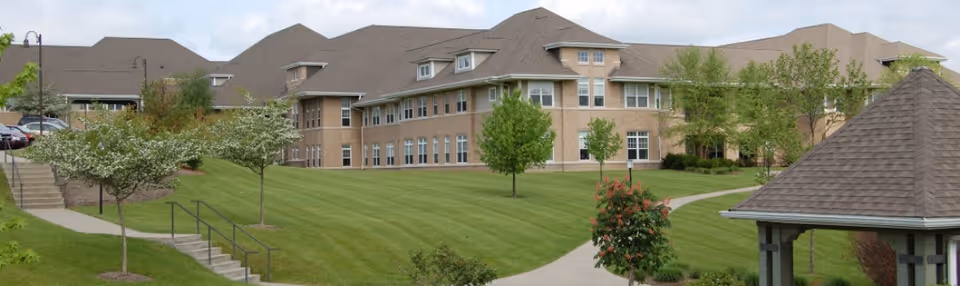 Exterior view of a senior living facility named Concordia at Rebecca Residence, showing a large multi-story brick building with multiple windows, surrounded by well-maintained green lawns, trees, and paved walkways. There is a gazebo structure on the right side and a parking area partially visible on the left.