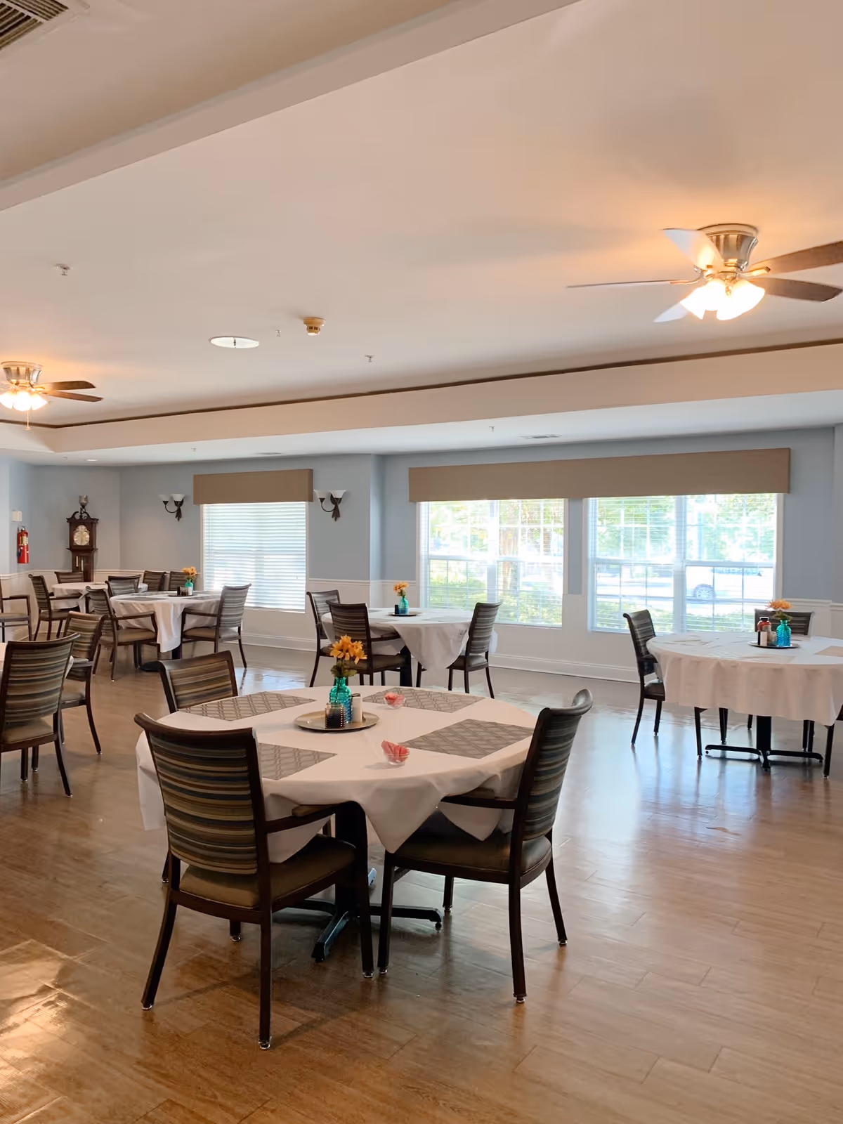 A bright dining room with several round tables covered with white tablecloths and gray placemats. Each table has a small vase with yellow flowers. The room has large windows with beige valances, wooden flooring, ceiling fans with lights, and wall sconces. Chairs with striped upholstery surround the tables.