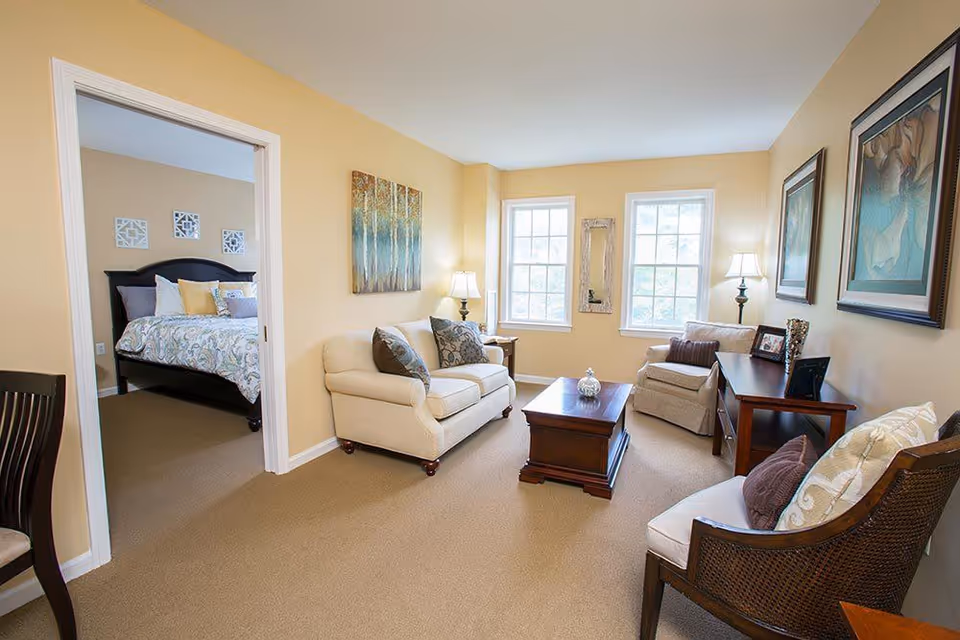 A cozy living room with beige walls and carpet, featuring a cream-colored sofa with patterned pillows, a wooden coffee table, an armchair, and a side table with a lamp. Two large windows let in natural light. Adjacent to the living room is a bedroom with a bed, pillows, and decorative wall art.