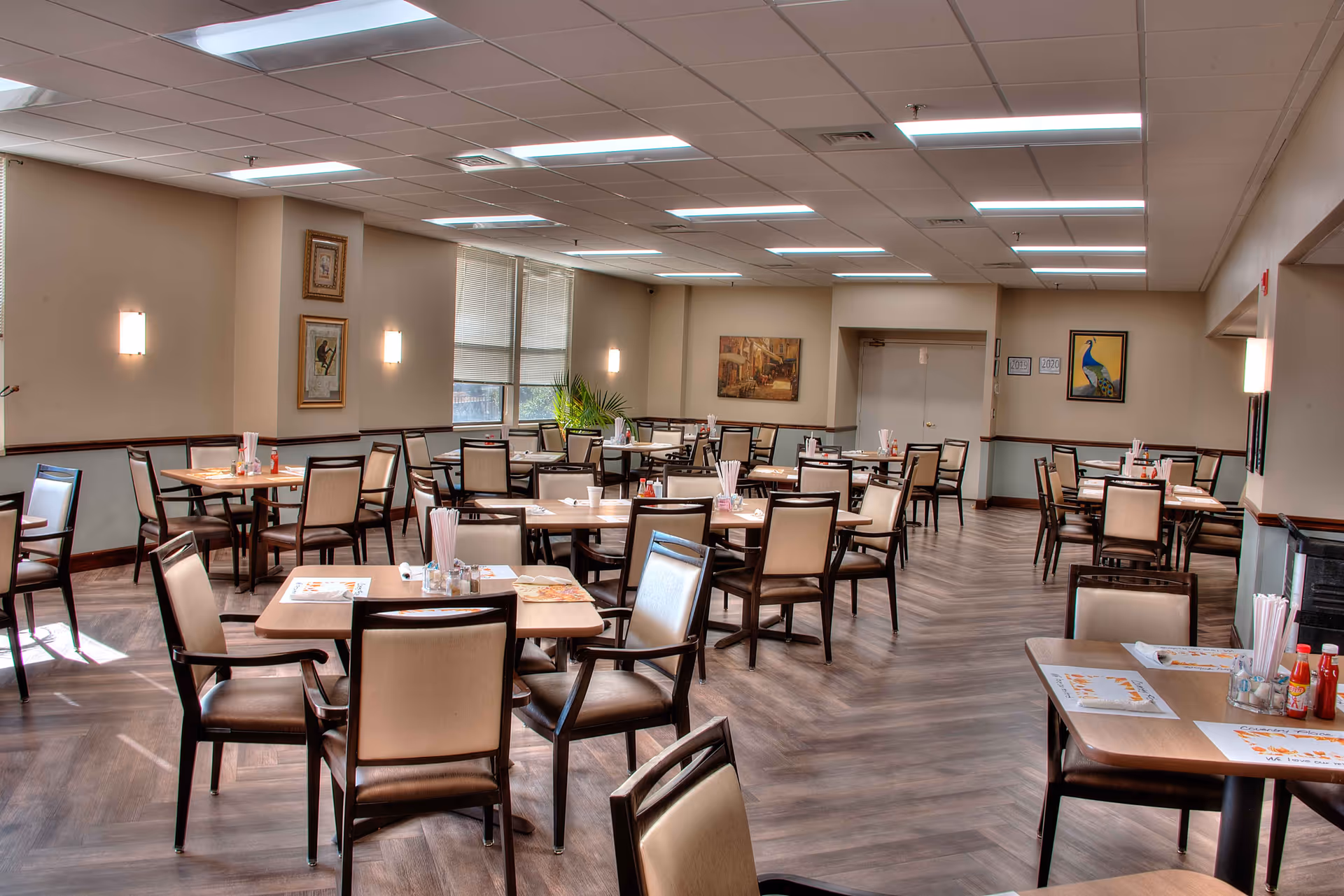 A spacious dining room with multiple wooden tables and chairs arranged neatly. Each table has placemats, napkins, condiments, and straws. The room has large windows with blinds, framed artwork on the walls, and bright overhead lighting. The floor has a wood-like finish.