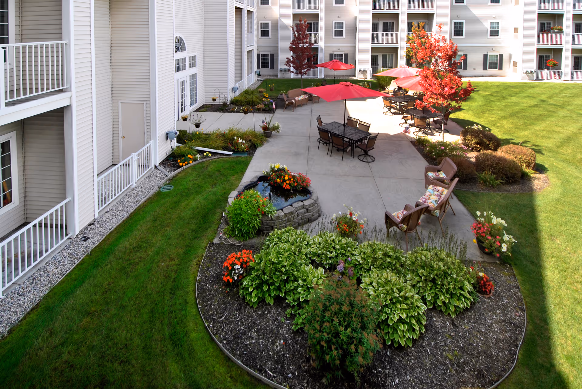Outdoor courtyard area of a retirement community with green grass, flower beds, and a concrete patio. The patio has tables with red umbrellas, chairs, and benches. The surrounding building has balconies and white siding. Trees with red leaves and various plants add color to the scene.