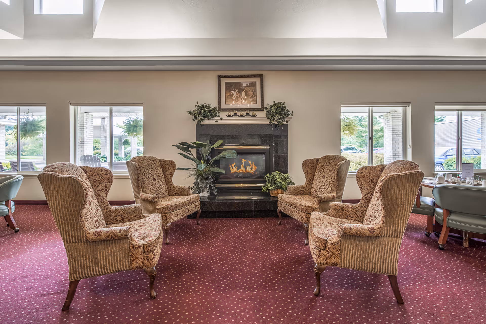 Cozy lounge with six patterned wingback chairs arranged around a lit fireplace beneath skylights.