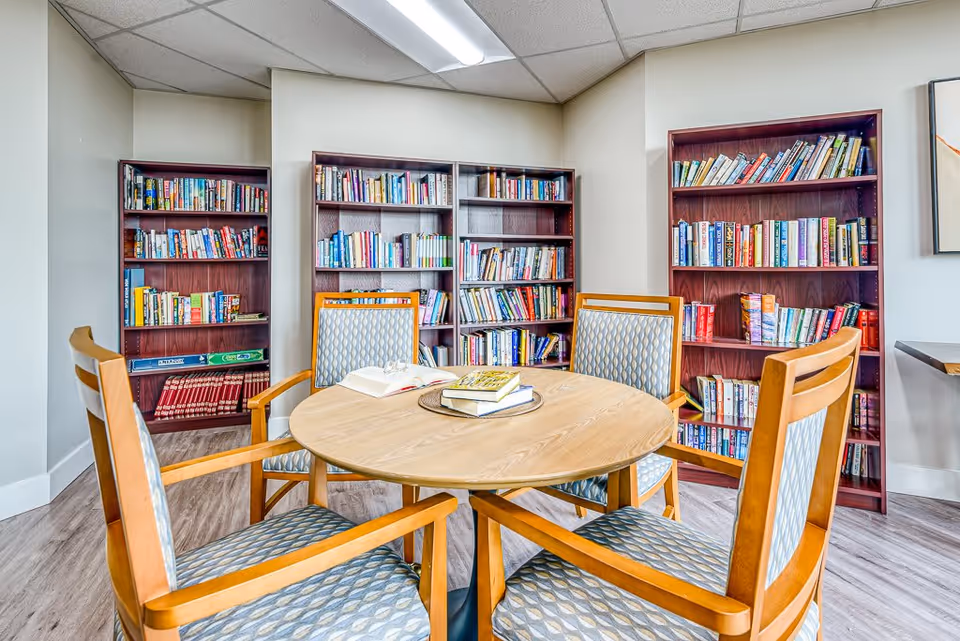A cozy reading area in a senior living facility featuring a round wooden table surrounded by four wooden chairs with patterned cushions. Behind the table are three wooden bookshelves filled with various books. The room has light-colored walls and a wood-patterned floor.