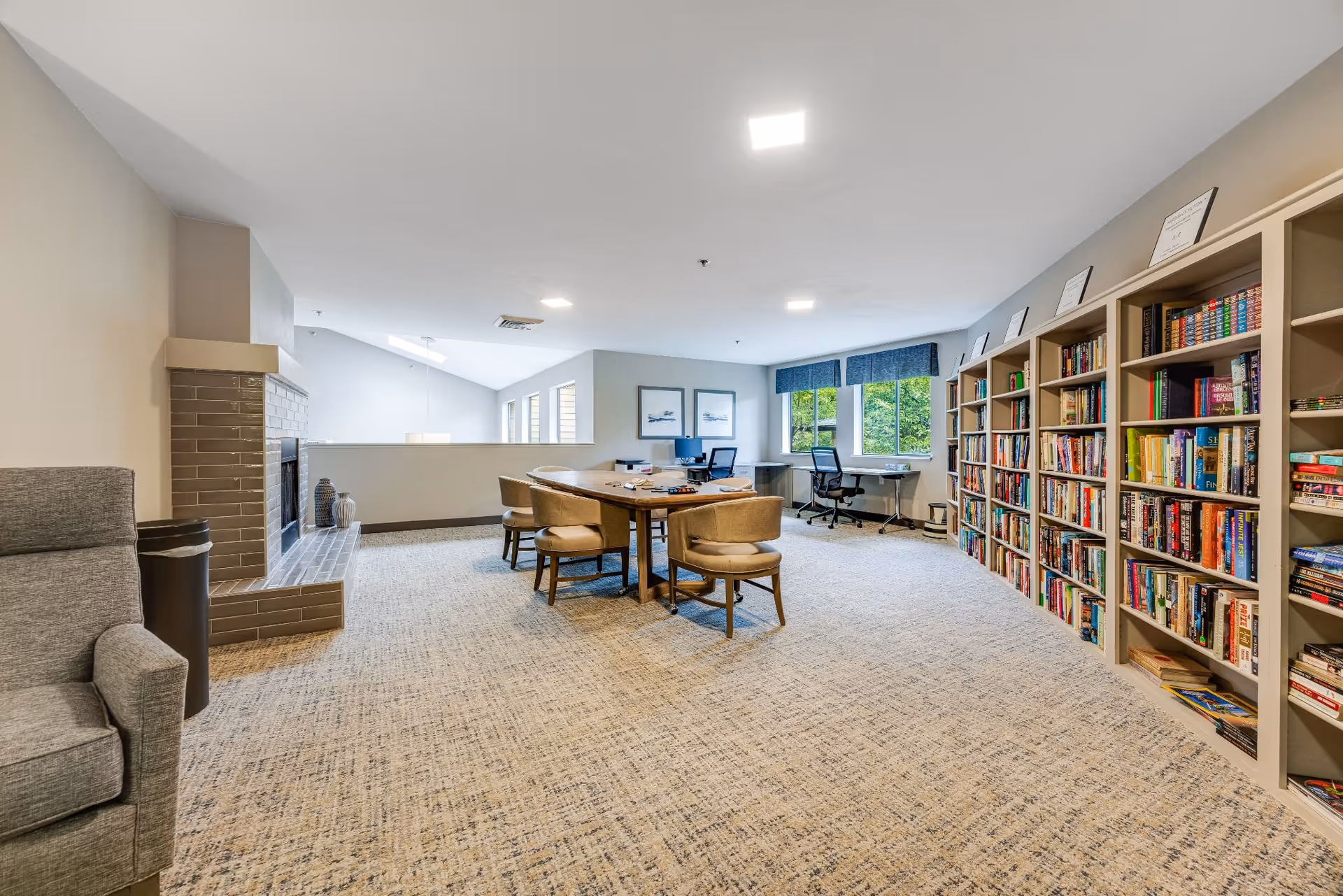 A spacious, well-lit room with a large wooden table surrounded by six cushioned chairs in the center. To the right, there is a long bookshelf filled with books and framed certificates on top. At the back, two desks with office chairs are positioned in front of windows with blue valances, showing greenery outside. On the left side, there is a gray armchair next to a fireplace with a tiled surround and decorative vases on the hearth. The room has beige carpet and light-colored walls.