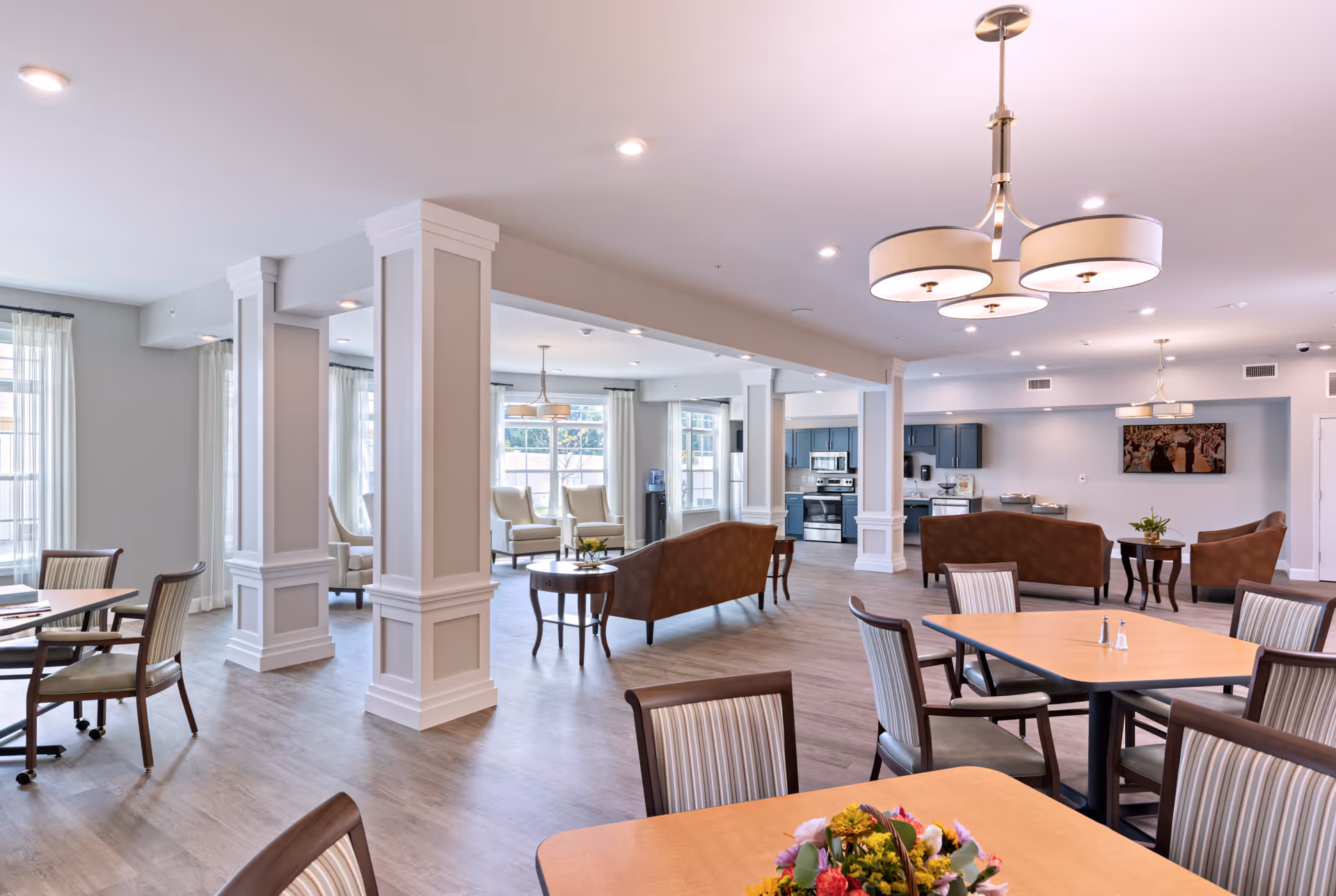 Bright open communal dining and lounge area with tables and chairs, upholstered sofas, decorative columns, and a kitchenette in the background.