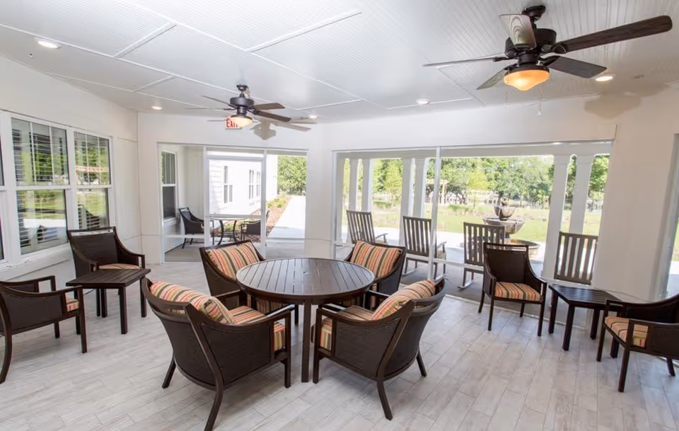 A bright indoor seating area with multiple wicker chairs featuring striped cushions arranged around a round wooden table. The room has large windows and glass doors that open to an outdoor patio with rocking chairs and a fountain visible outside. Ceiling fans with lights are mounted on the white paneled ceiling.