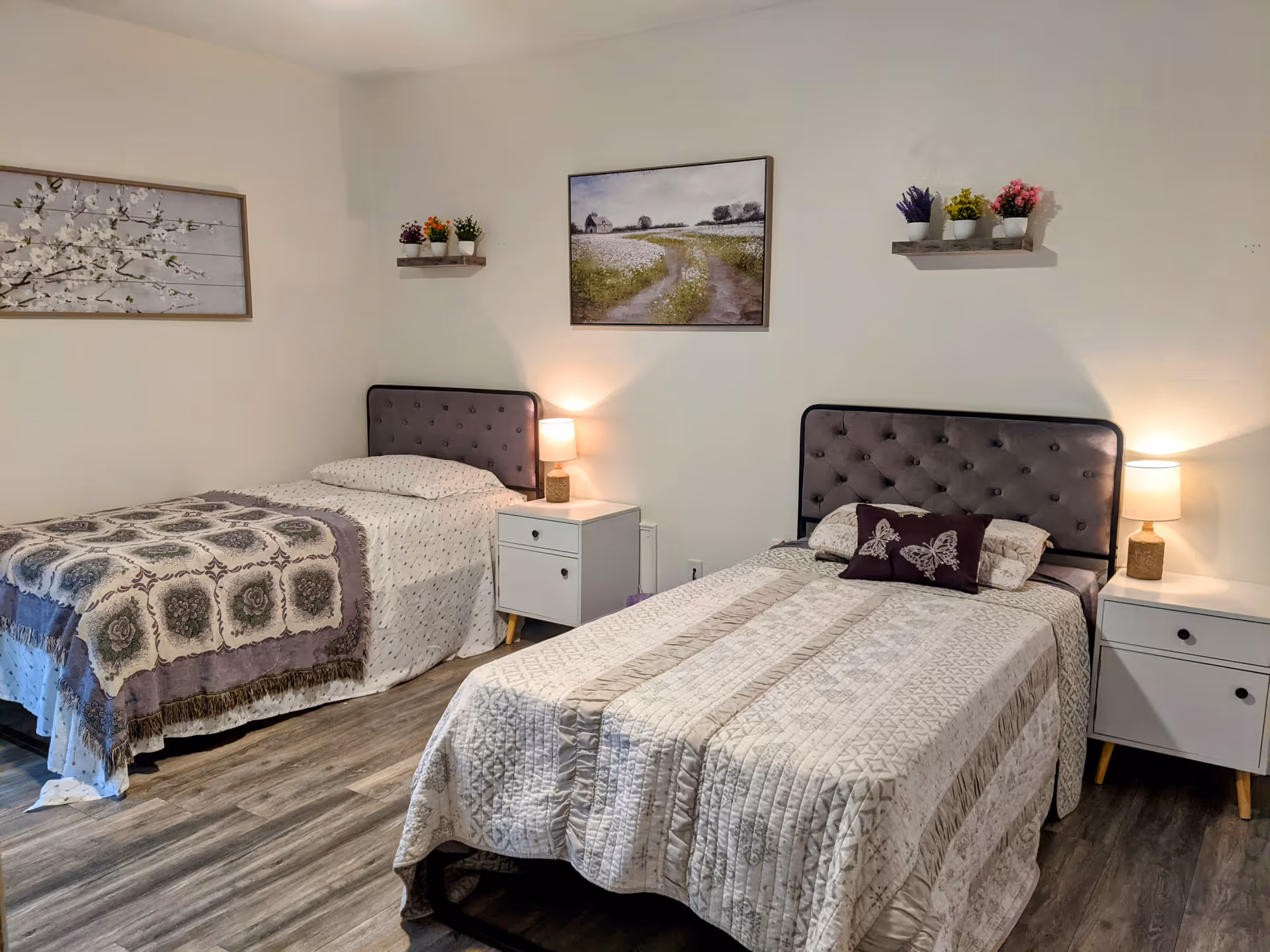 Two twin beds with tufted headboards, nightstands with lamps, wall art and potted plants in a tidy bedroom.