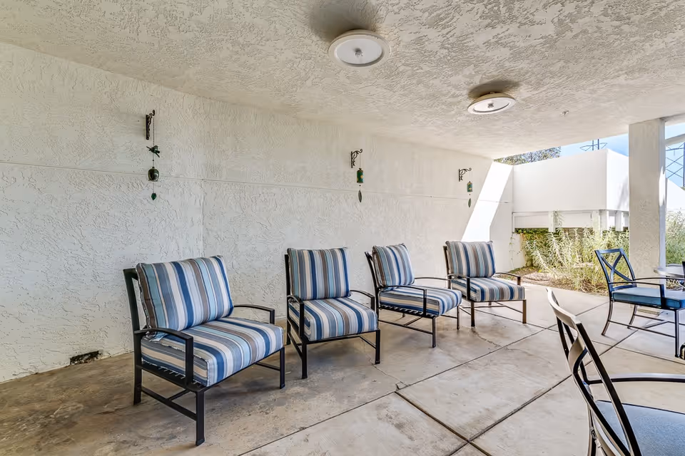 Covered outdoor seating area with four striped cushioned chairs arranged in a row against a textured white wall, and additional metal chairs and tables nearby. The space has a concrete floor and ceiling with mounted light fixtures, and some greenery visible outside.
