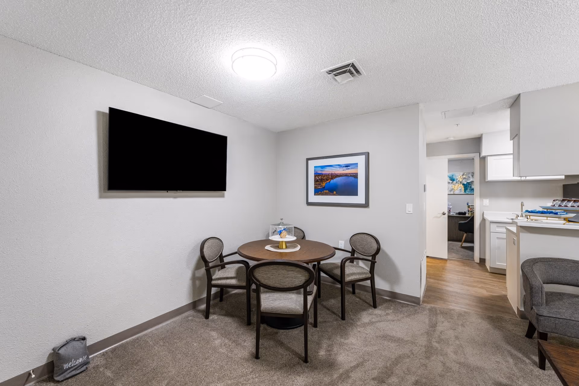 A small dining area with a round wooden table surrounded by four cushioned chairs. On the table is a cake stand with a glass cover. A flat-screen TV is mounted on the wall to the left, and a framed picture of a lake scene hangs on the wall behind the table. To the right, there is a partial view of a kitchen with white cabinets and a sink, and an open doorway leading to another room. The floor is carpeted in the dining area and transitions to wood flooring in the kitchen.