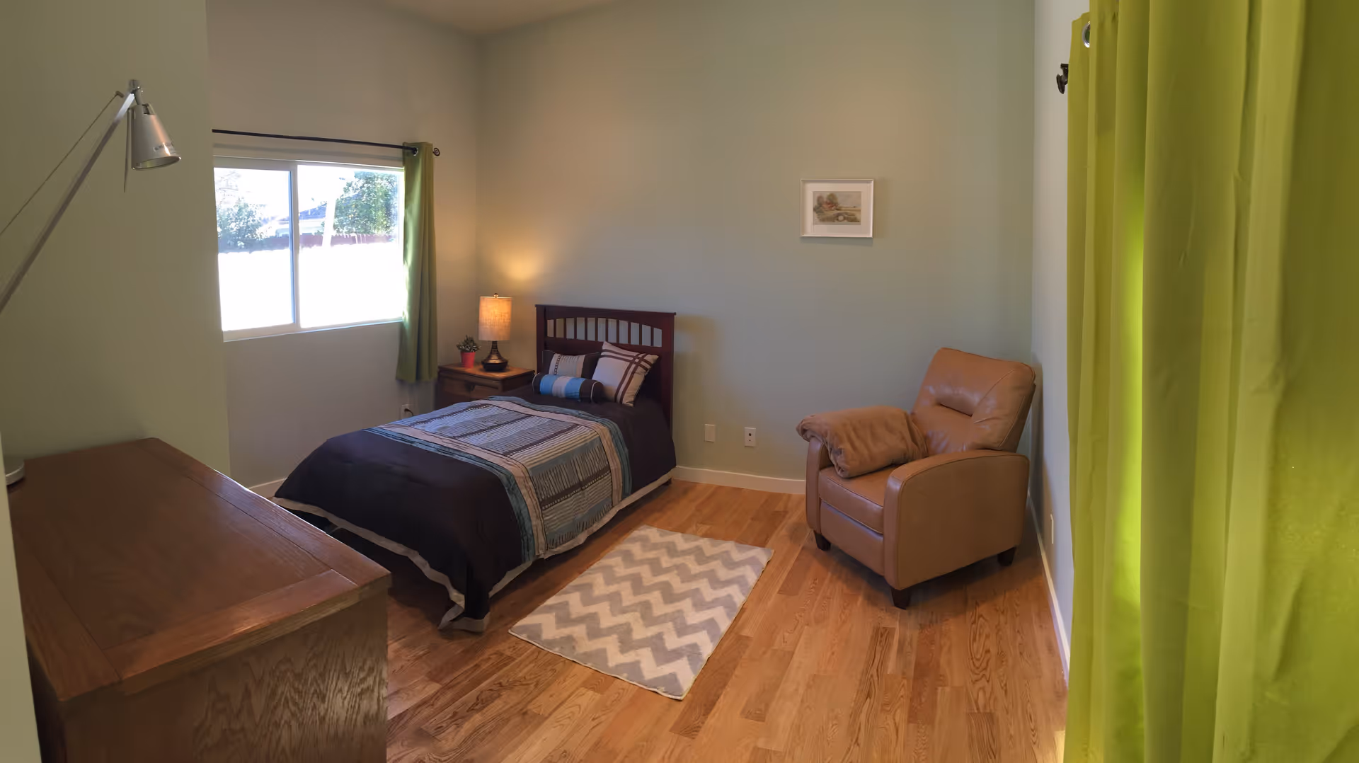 A small bedroom with light green walls and wooden flooring. The room contains a single bed with a dark wooden headboard, a blue and gray striped bedspread, and several pillows. Next to the bed is a wooden nightstand with a lamp and a small plant. There is a large window with green curtains letting in natural light. A beige armchair is placed against the right wall, and a wooden dresser is on the left side of the room. A small chevron-patterned rug is on the floor near the bed.