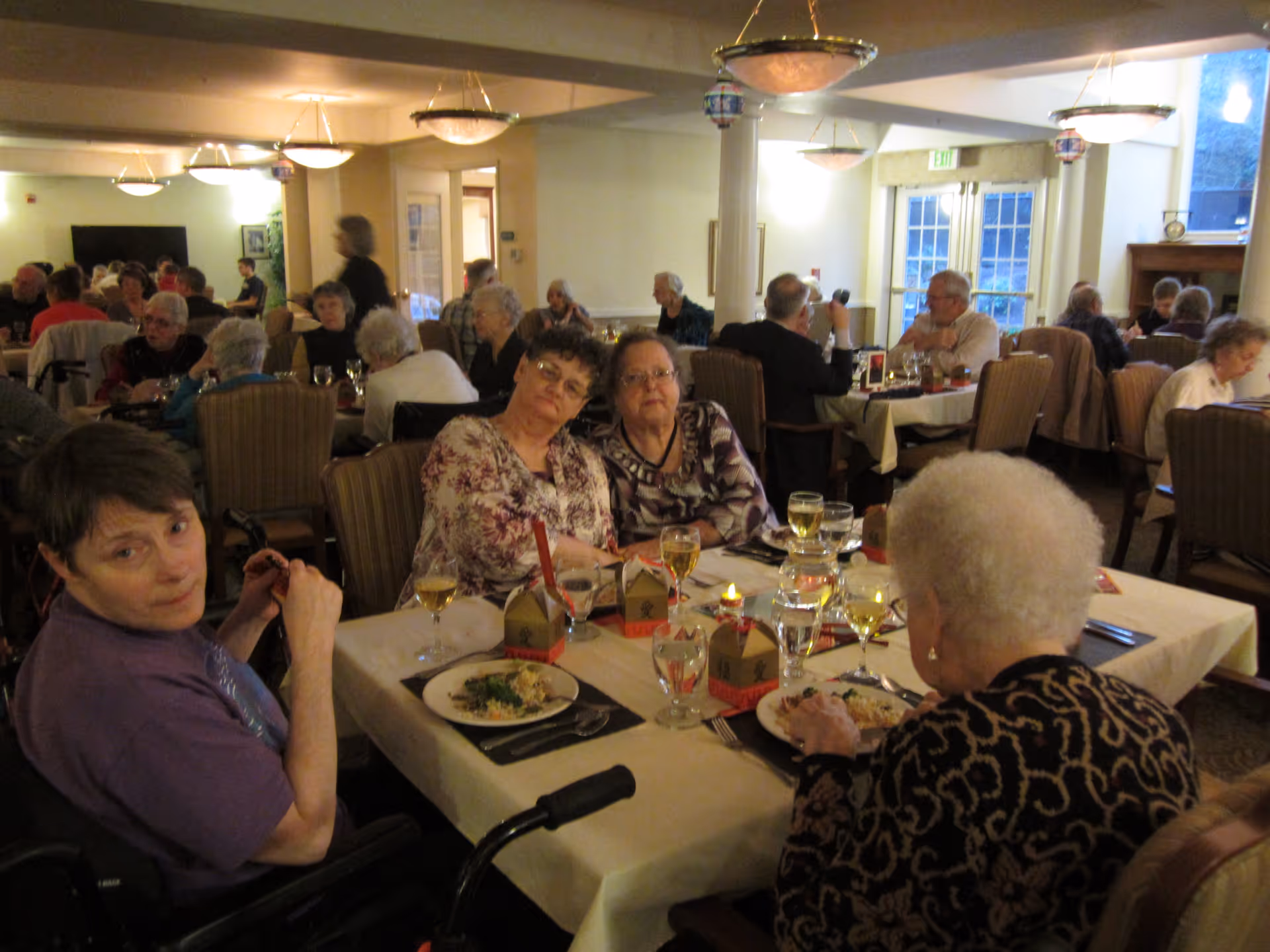 A dining room in a senior living facility with several elderly people seated at tables enjoying a meal. The room is warmly lit with hanging light fixtures, and tables are set with plates of food, glasses of water and wine, and small decorative items. The atmosphere appears social and communal.