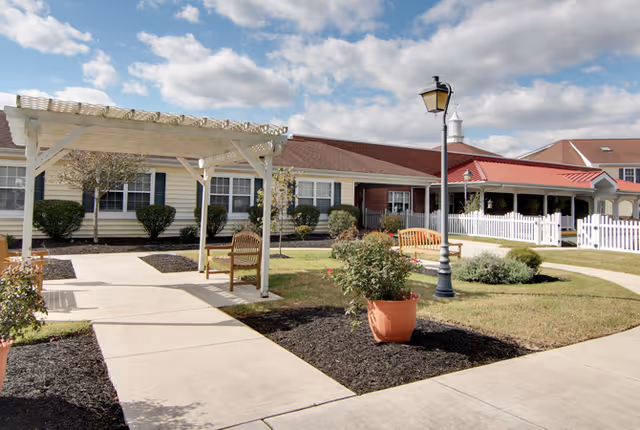 Outdoor courtyard with a white pergola, wooden benches, a lamppost, potted plants, and walkways in front of the Milford Place building.