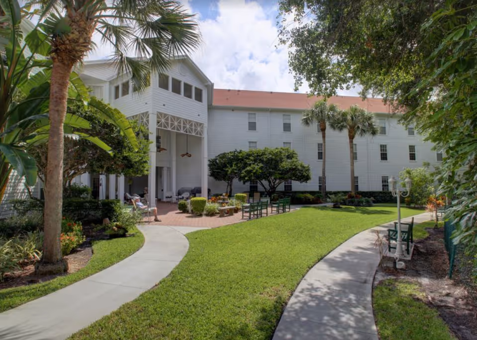Courtyard with paved walkways, green lawn, benches and palm trees in front of a white multi-story senior living building.