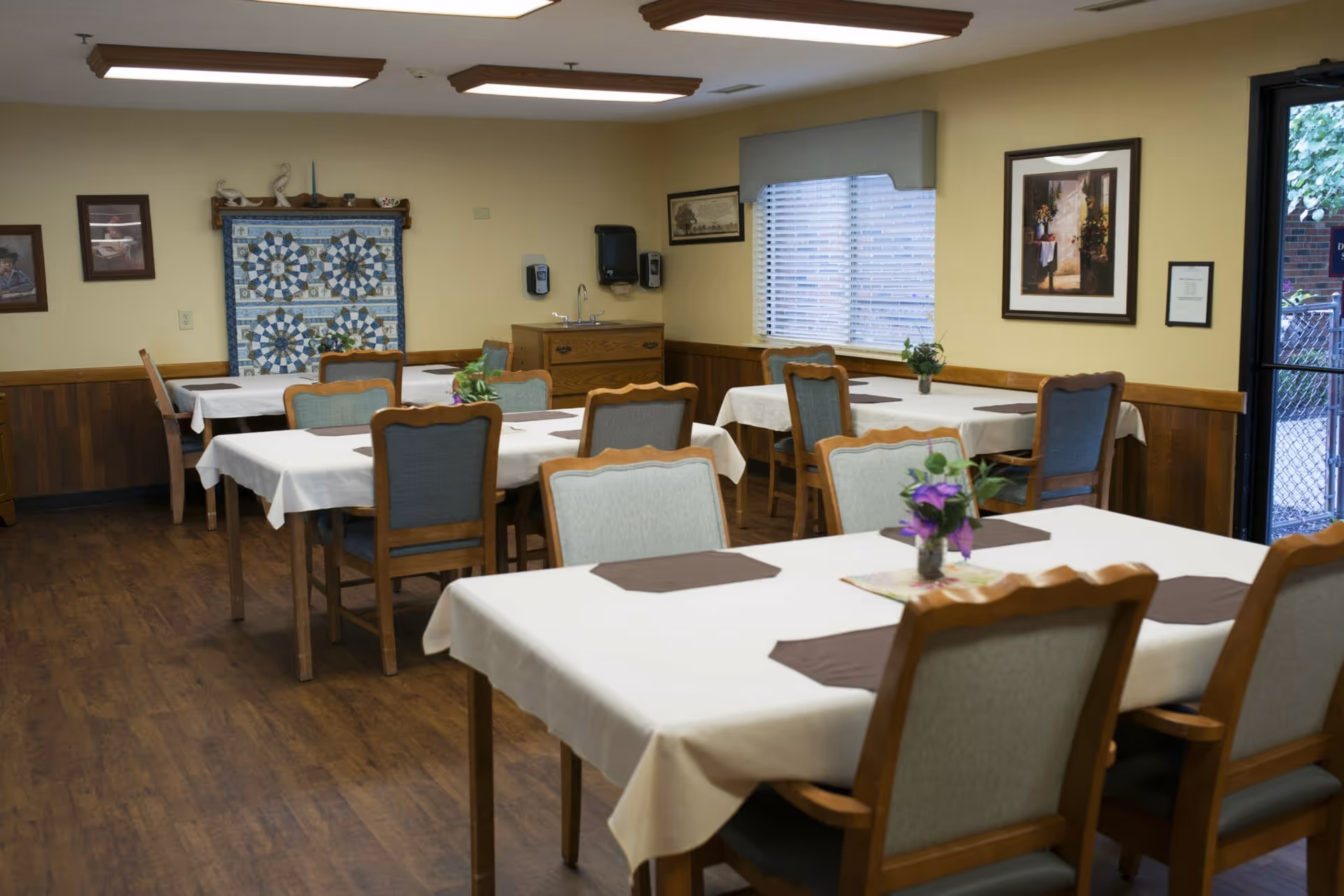 A dining room with several tables covered in white tablecloths and brown placemats. Each table has a small vase with purple flowers. The room has wooden floors and wood paneling on the lower half of the walls. There are framed pictures and a quilt hanging on the walls. A window with blinds and a door leading outside are visible.