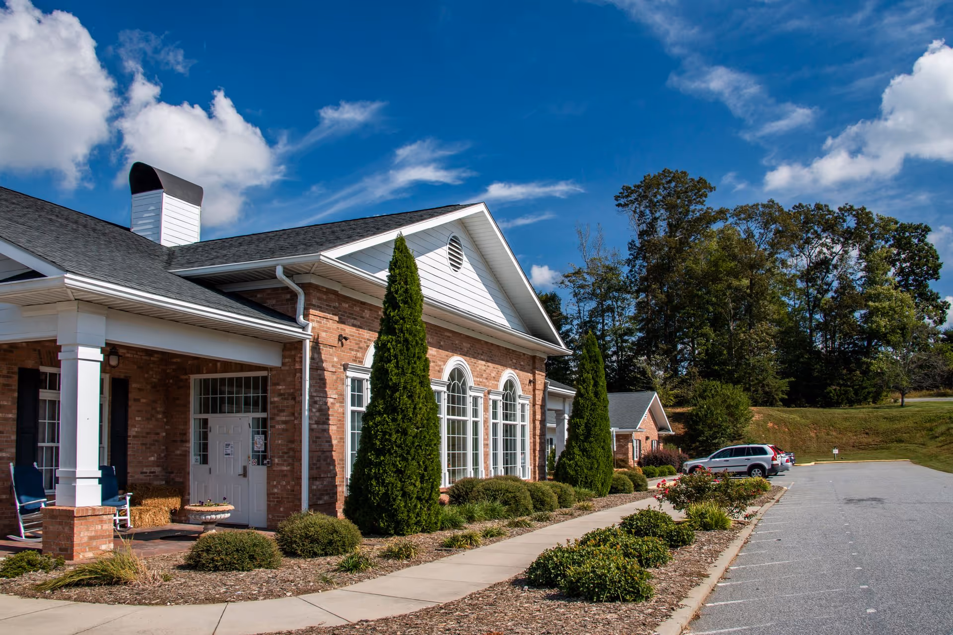 Exterior view of a brick building with white trim under a blue sky with some clouds. The building has large arched windows and is surrounded by landscaped bushes and trees. There is a sidewalk and a parking area with a few parked cars.