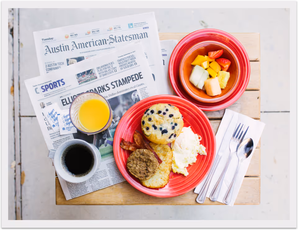 Overhead view of a breakfast on a wooden table with coffee, orange juice, a red plate of eggs, muffin, bacon and sausage, a bowl of fruit, utensils on a napkin, and newspapers.