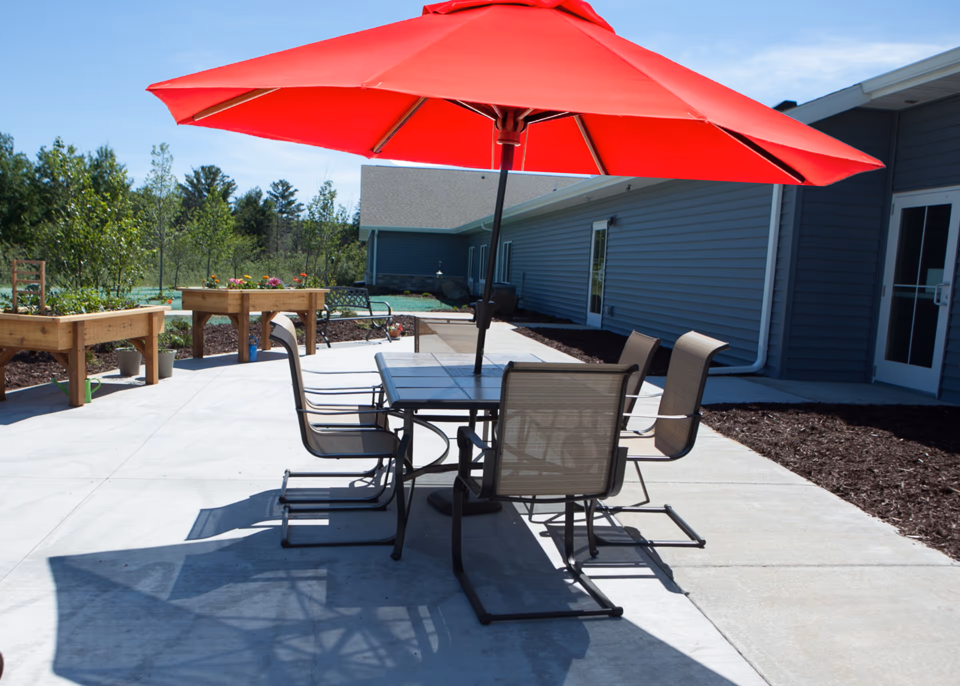 Outdoor concrete patio with a table, several chairs, and a large red umbrella beside a single-story building.