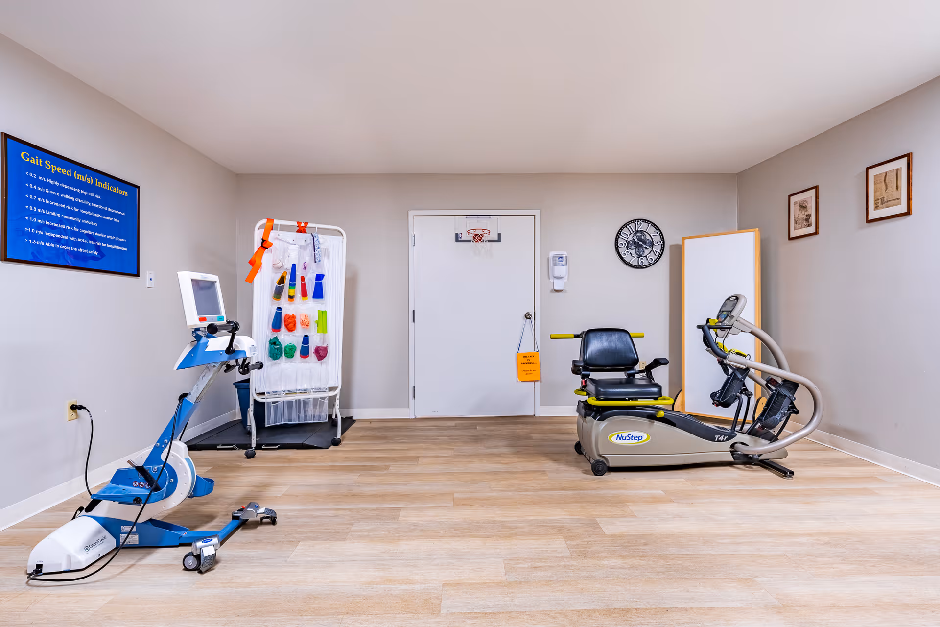 A rehabilitation or physical therapy room with exercise equipment including a stationary bike and a NuStep recumbent cross trainer. There is a chart on the wall titled 'Gait Speed (m/s) Indicators', a rack with colorful therapy bands, a wall clock, and framed pictures on the walls. The room has light wood flooring and neutral-colored walls.