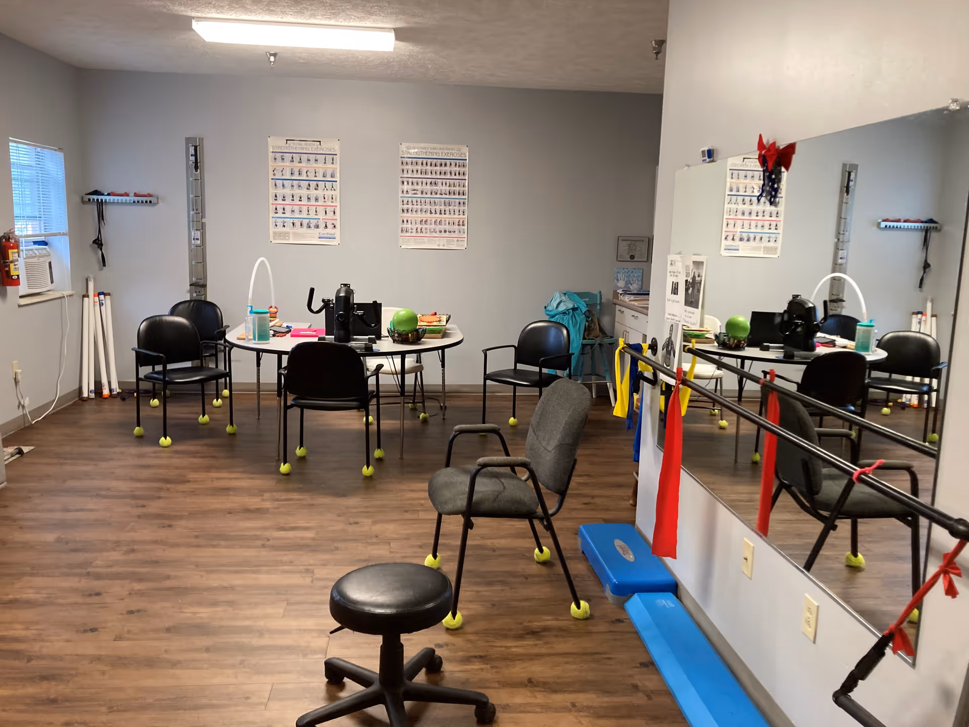 A small exercise room with wooden flooring and gray walls. There are several black chairs with tennis balls on the legs, a round table with exercise equipment and supplies, and a large mirror on one wall with resistance bands hanging on a ballet barre. Exercise posters are displayed on the walls.