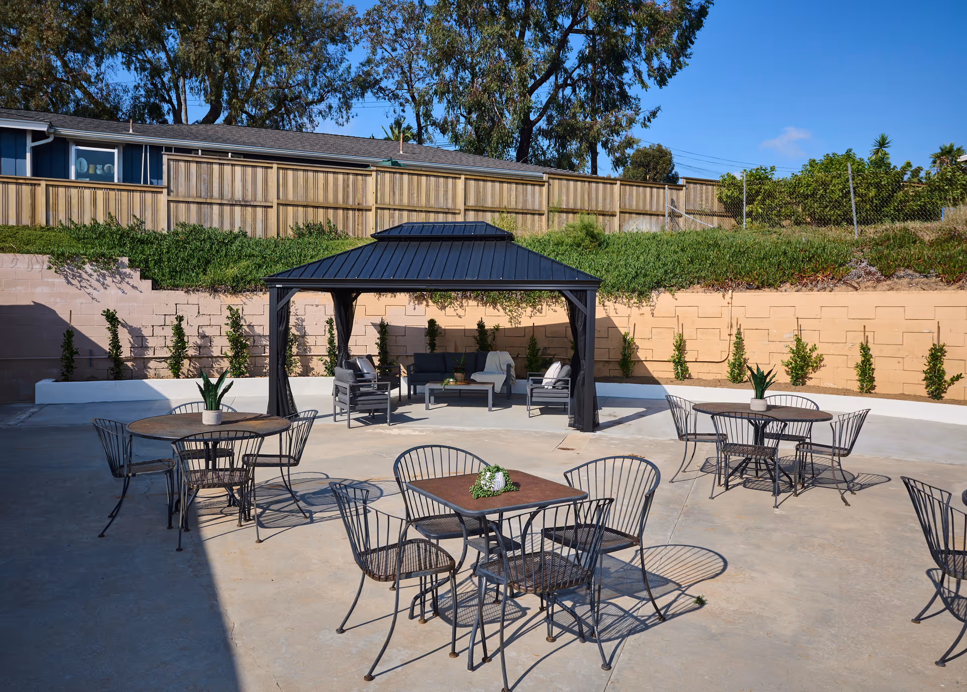 Outdoor patio area with several metal tables and chairs arranged on a concrete surface. A black gazebo with seating furniture underneath is positioned against a retaining wall with small plants growing along it. Trees and a wooden fence are visible in the background under a clear blue sky.