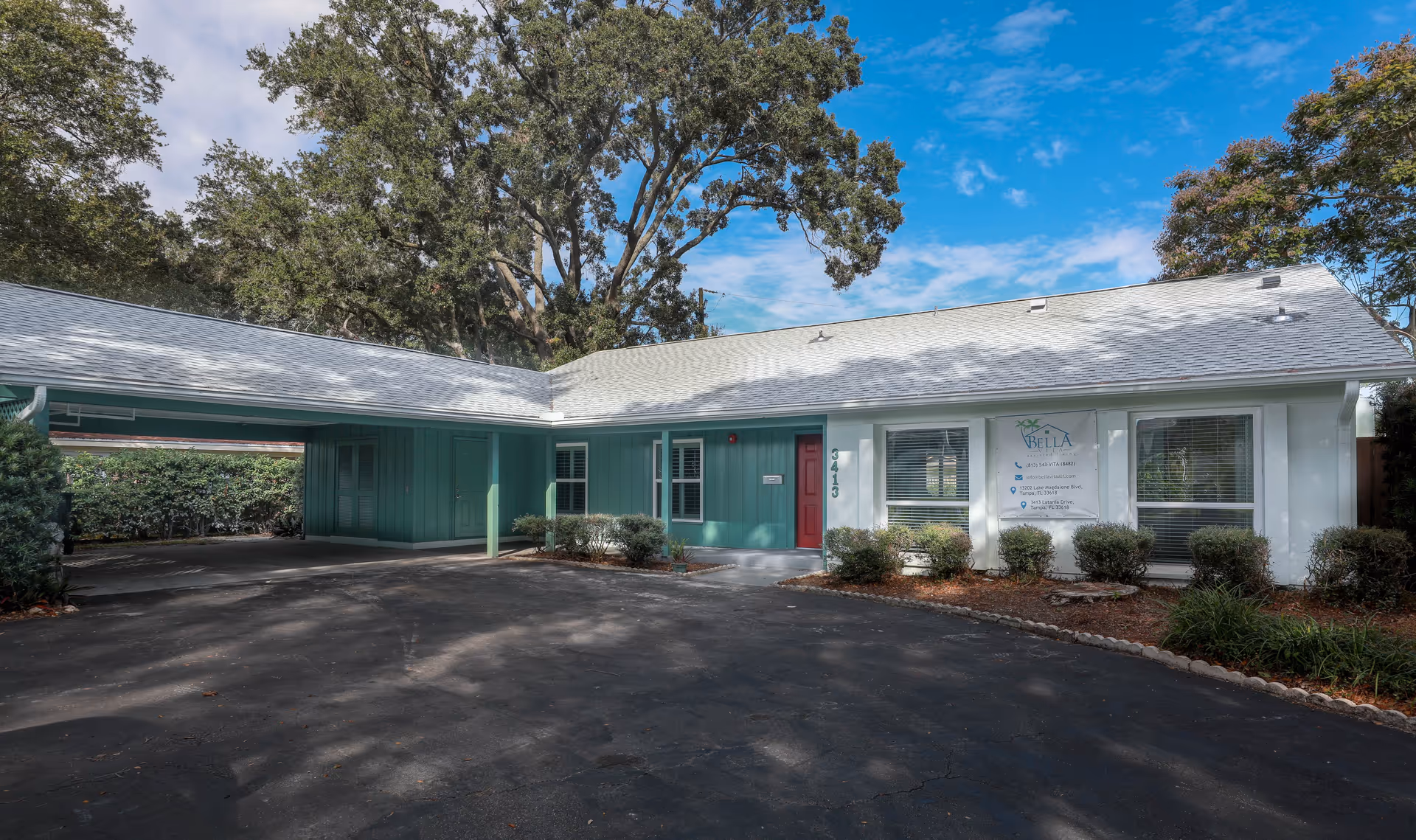 Exterior view of BELLA VITA ALF at LATANIA facility showing a single-story building with a white roof, green and white walls, a red door, and surrounding bushes and trees under a partly cloudy blue sky.
