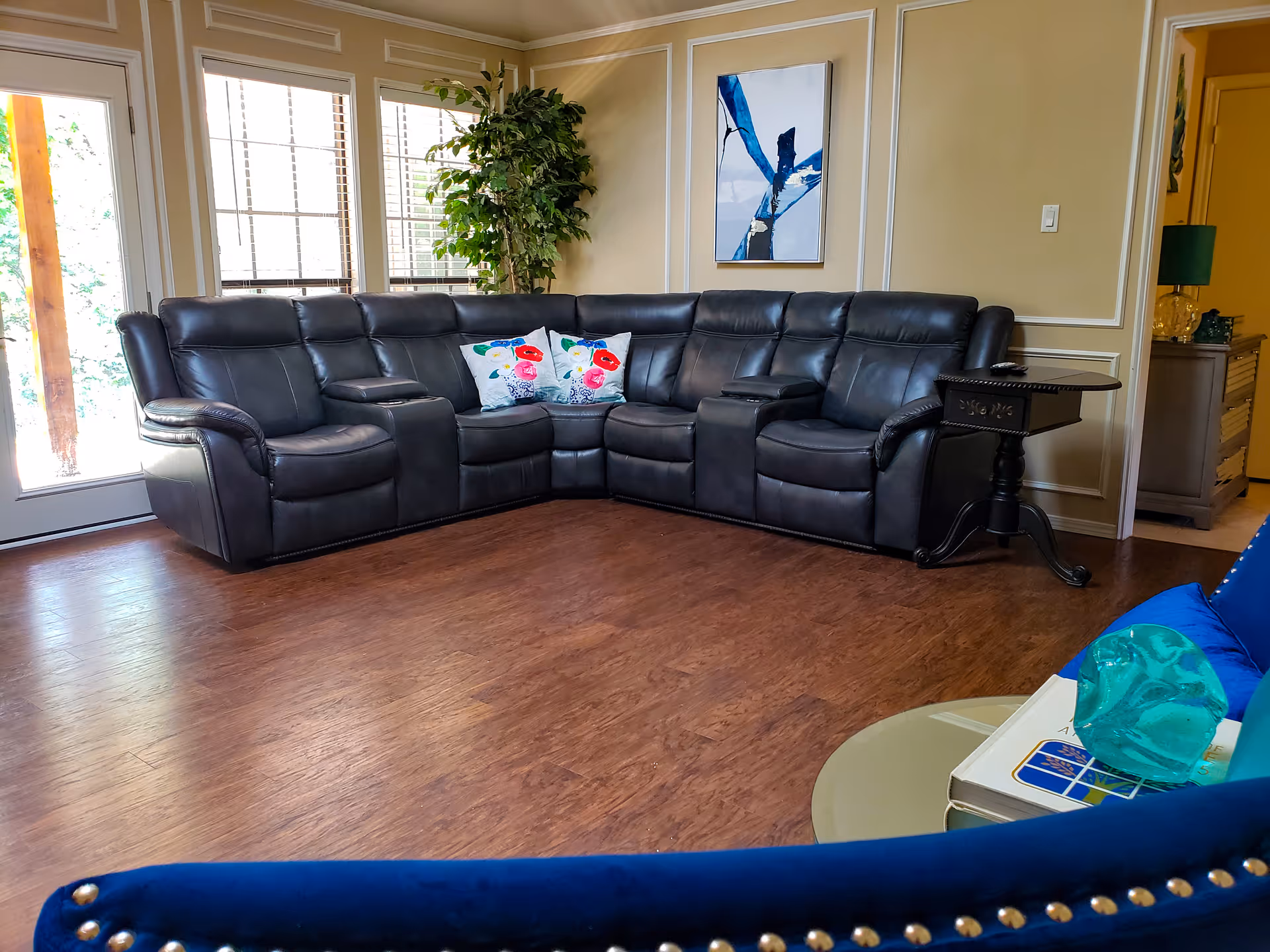 A living room with a large black leather sectional sofa adorned with two floral pillows. There is a small black side table next to the sofa and a green leafy plant in the corner near large windows with blinds. The walls are beige with decorative molding and a modern abstract painting. The floor is wooden, and part of a blue chair with nailhead trim is visible in the foreground along with a round table holding books and a decorative glass object.