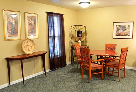 A small dining area with a round wooden table and four matching chairs with red cushions. The room has beige walls, a green patterned carpet, and a window with dark curtains. There is a narrow wooden console table against one wall with a decorative plate and two framed floral pictures above it. A metal shelving unit with decorative items is in the corner, and a framed picture hangs on the opposite wall.