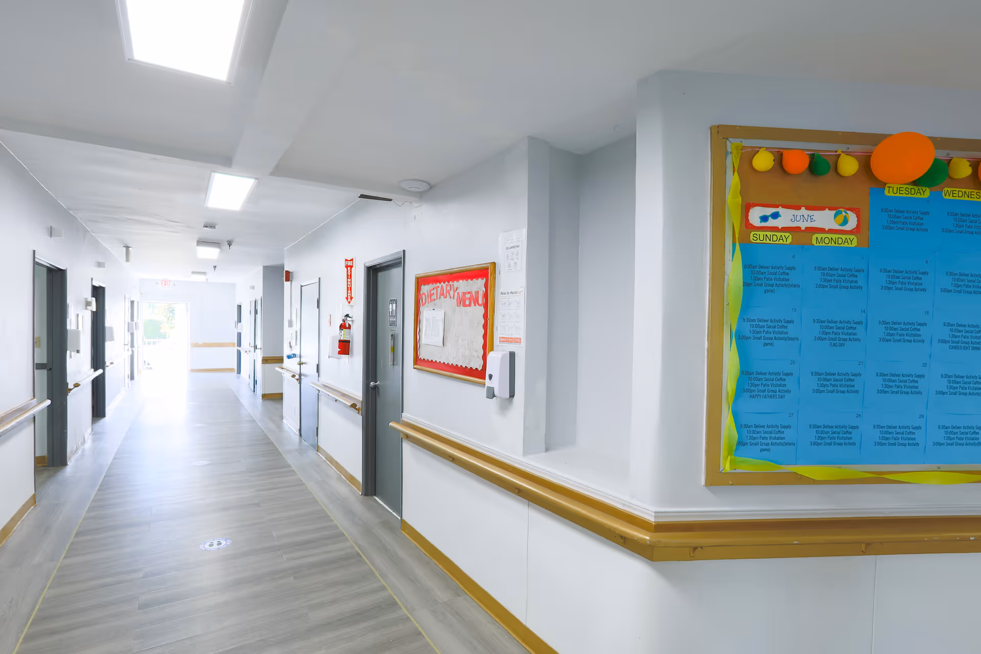 Bright, wide healthcare hallway with handrails, several doors, and a bulletin board on the right.