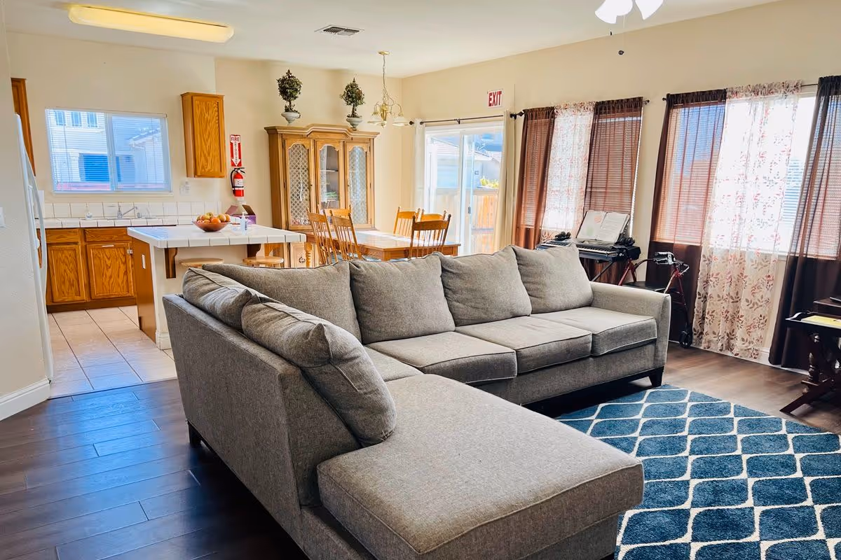 Open-plan living area with a gray L-shaped sectional sofa, blue patterned rug, dining table, and kitchen island.